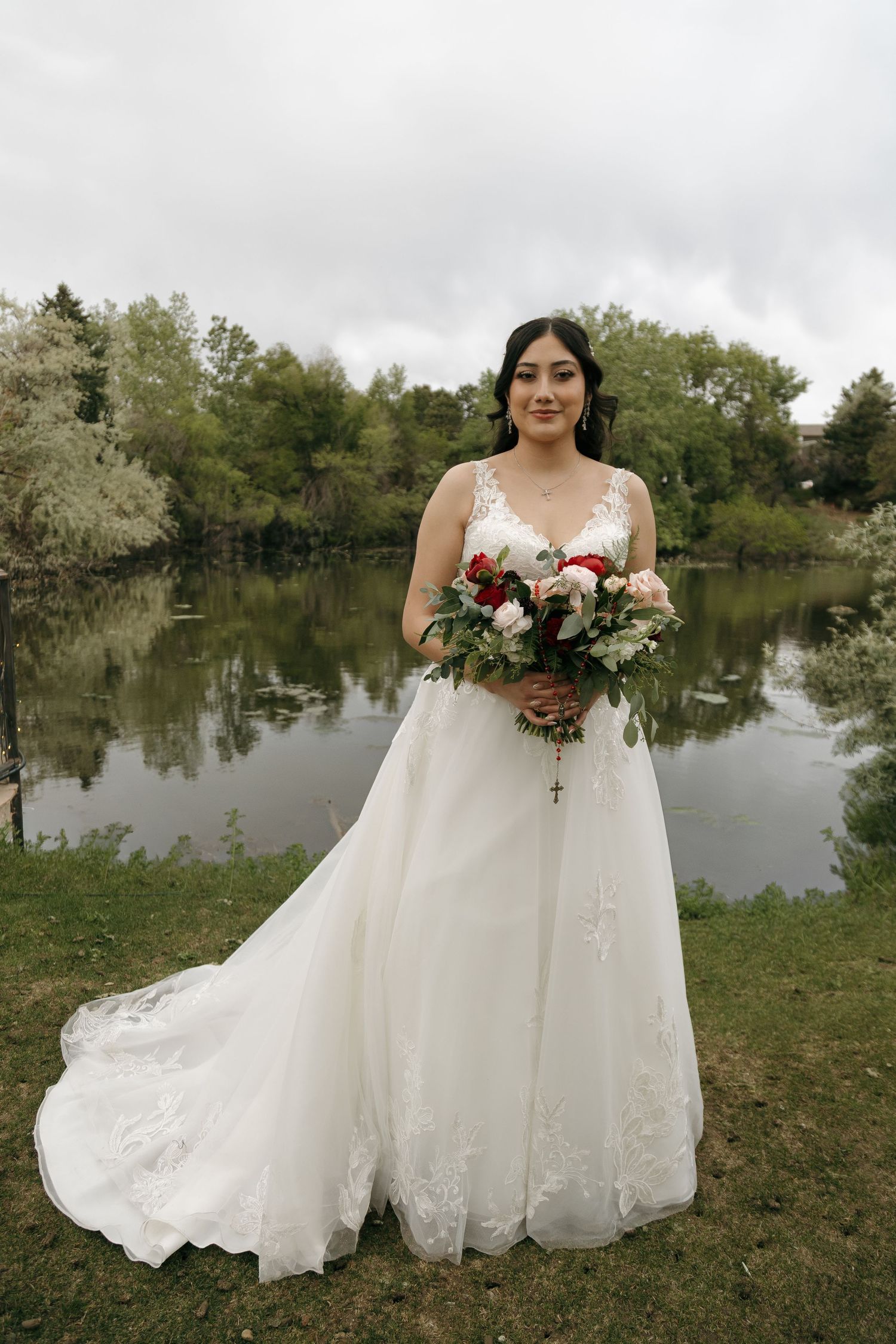 Bride in lace A-line wedding gown holding red and white bouquet by a pond.
