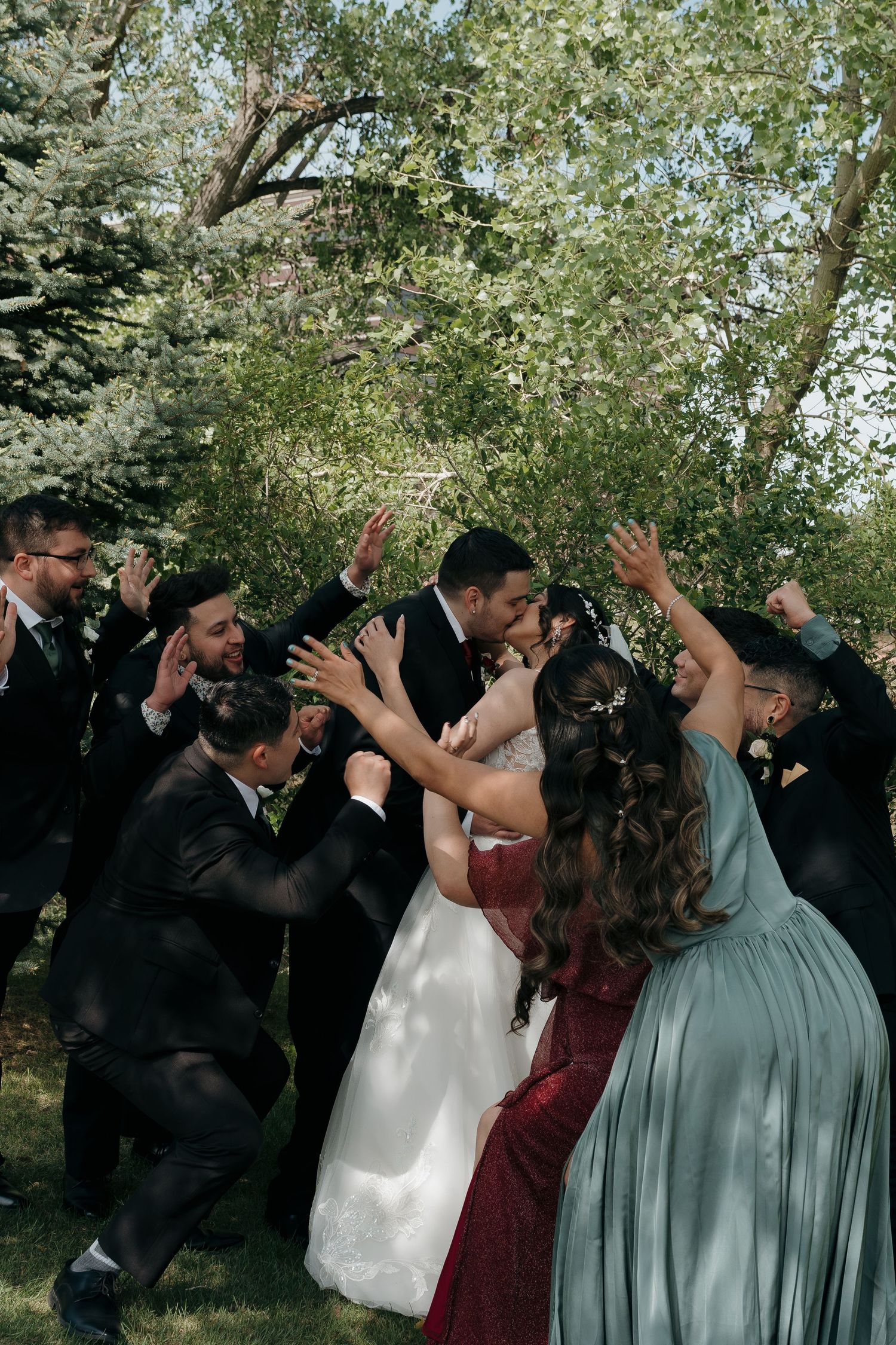 Bride and groom kissing as joyful wedding party members celebrate around them in a sunny outdoor setting.