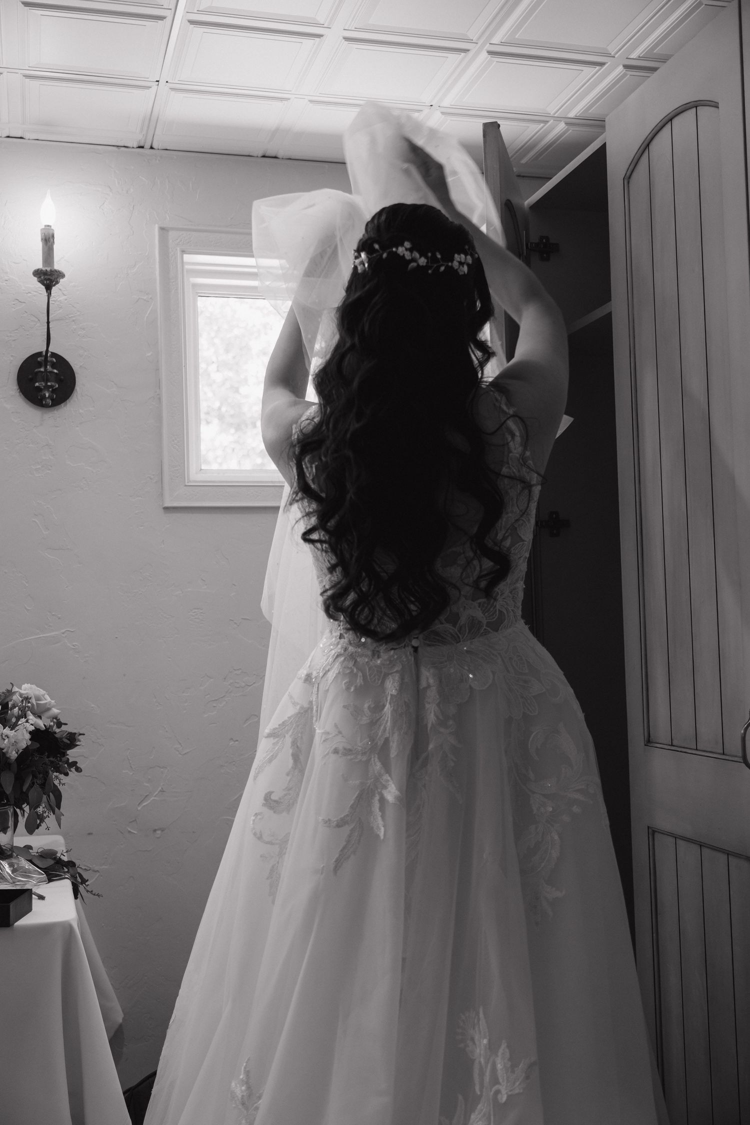 Bride in elegant lace wedding gown adjusting her veil indoors in black and white photo.