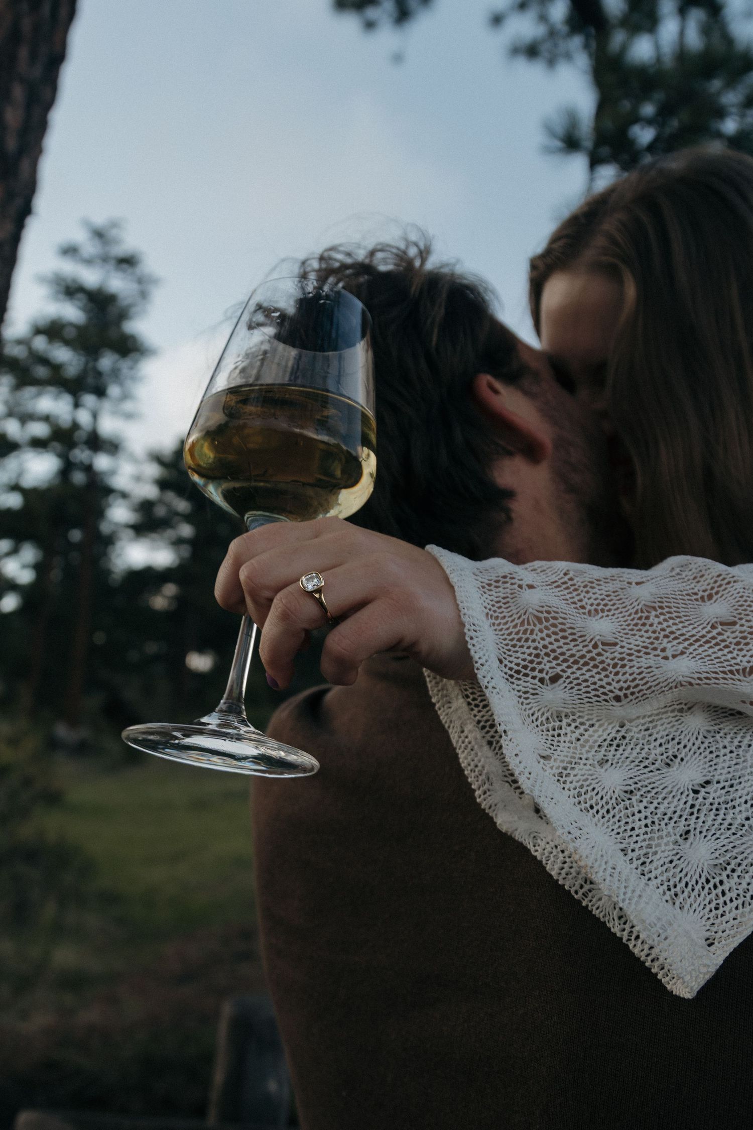 Woman holding a glass of white wine with an engagement ring while embracing her partner outdoors.