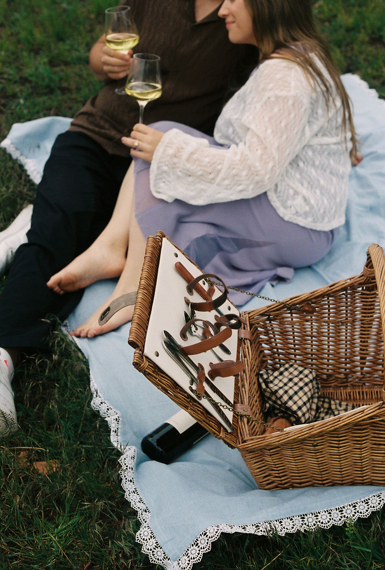 Close-up of a couple enjoying white wine on a lace-trimmed blanket beside a wicker picnic basket.