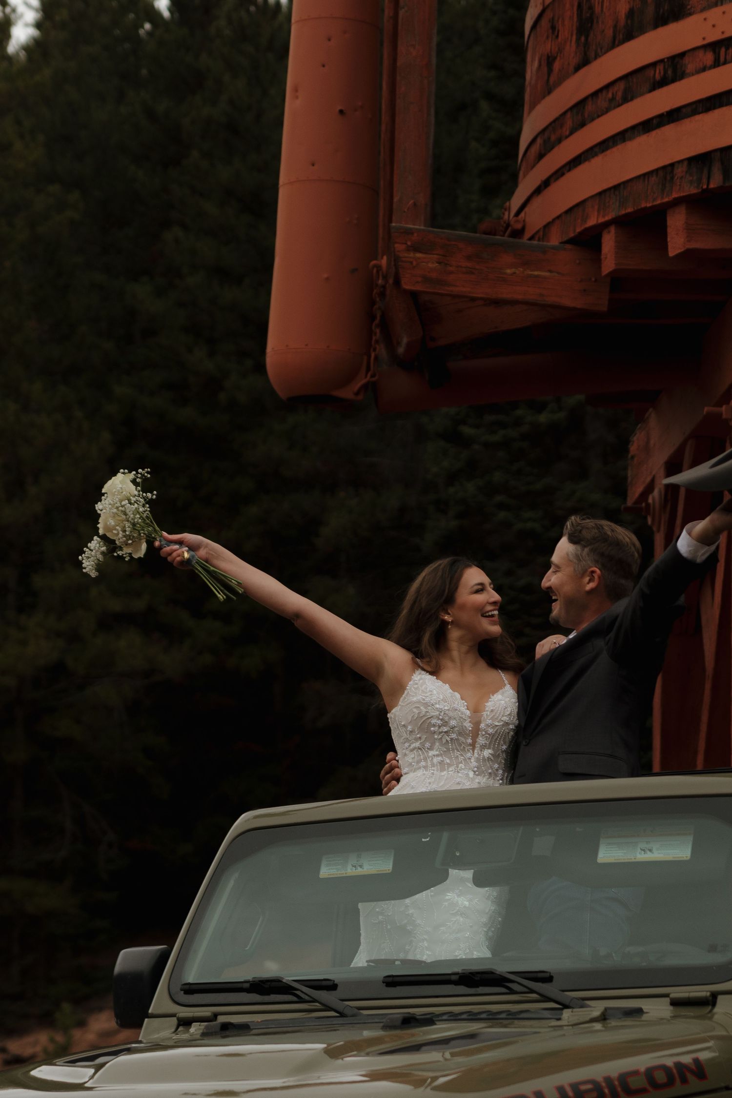 Newlyweds celebrate atop a Jeep with raised bouquet against rustic architecture.