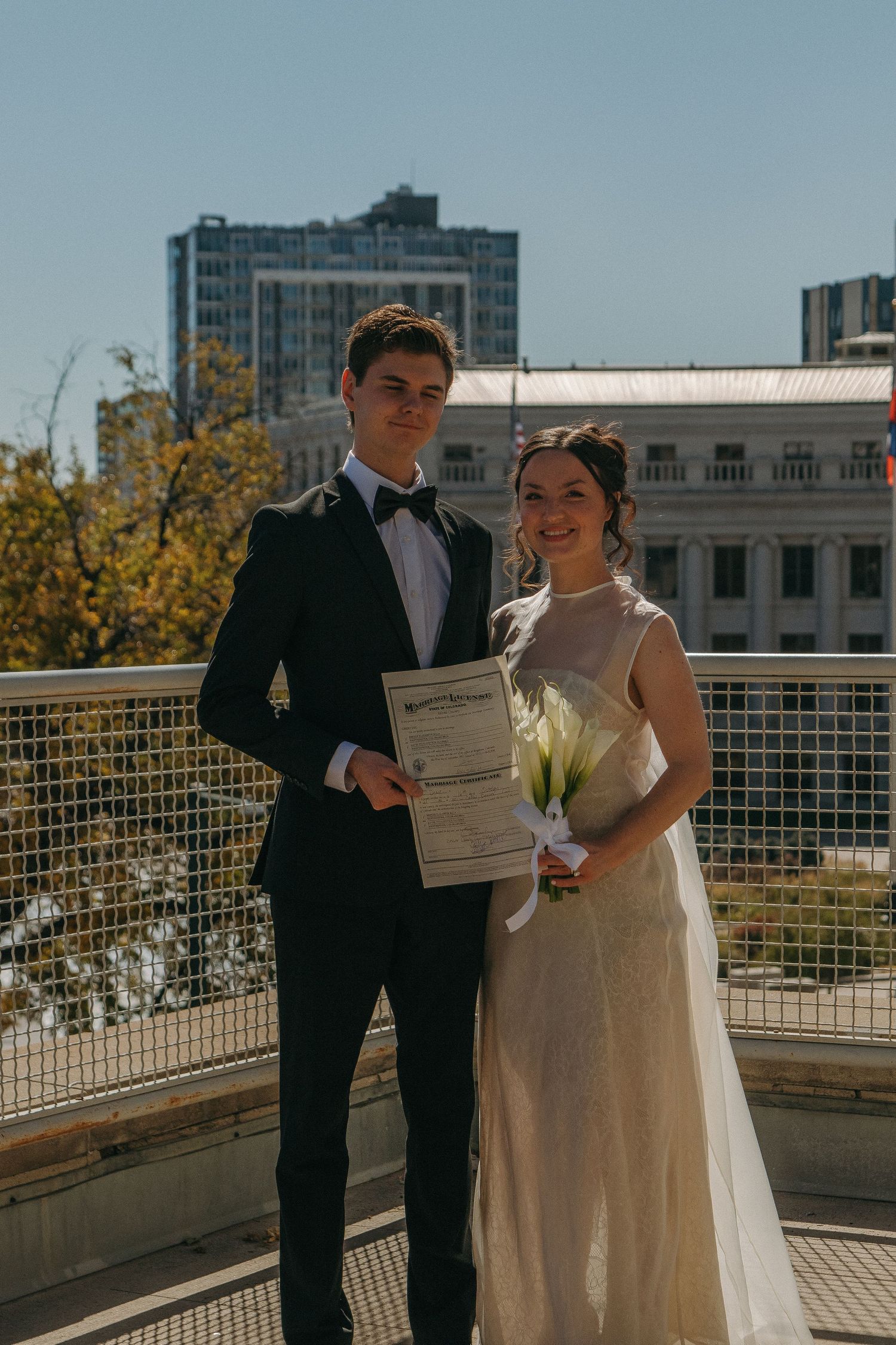 Wedding couple holding documents on a rooftop with city buildings in background.