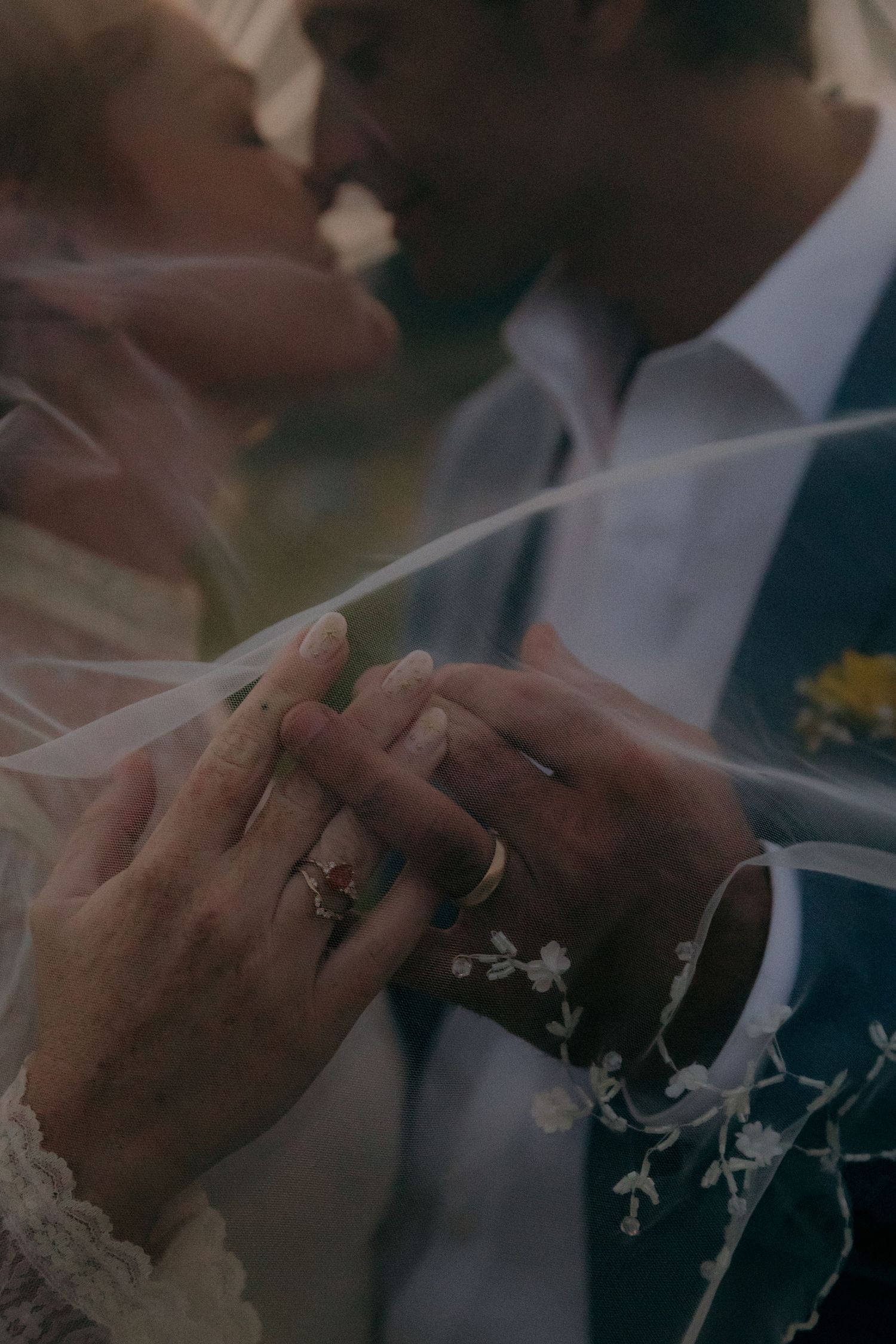 Close-up of bride and groom holding hands showing wedding rings under a flowing veil.