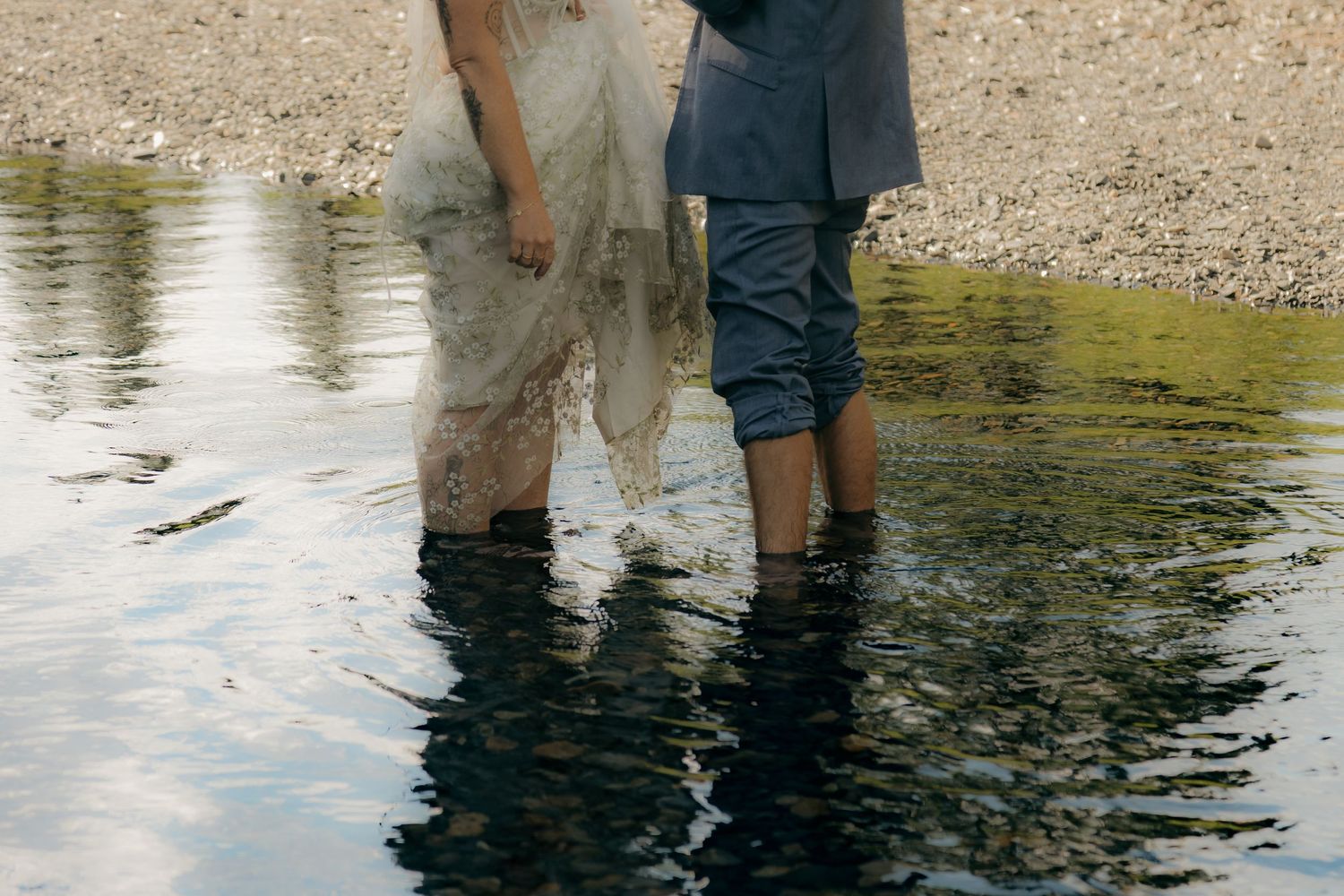 Couple standing in shallow water, wearing wedding attire, with rocky shoreline in background.