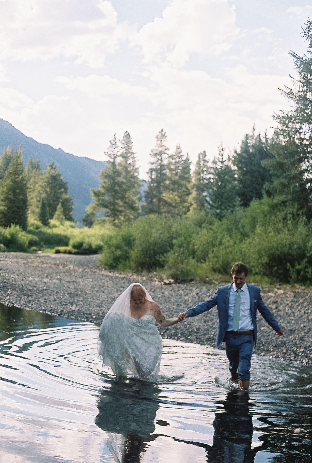 A bride and groom wade through a mountain river holding hands on their wedding day.