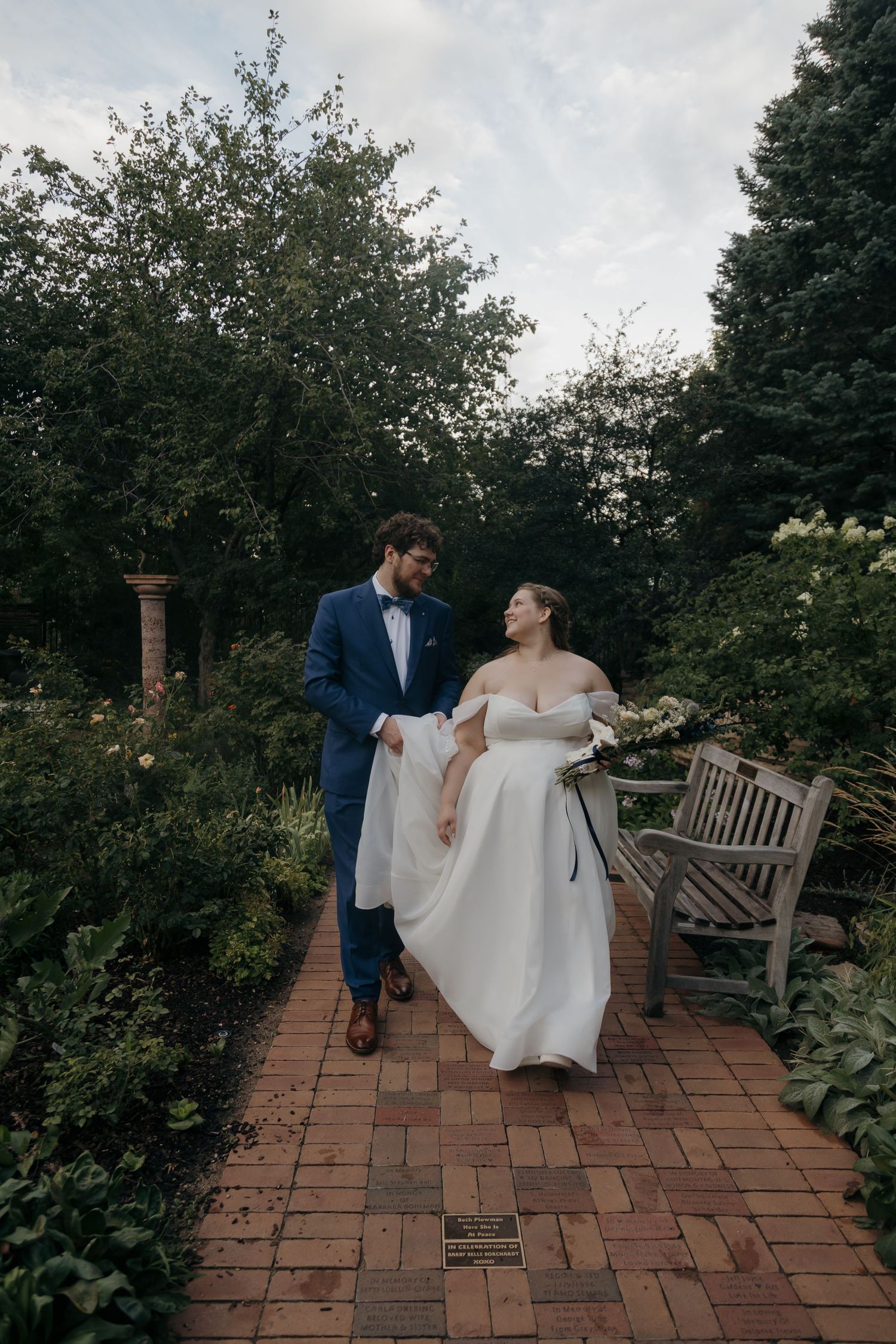 Bride and groom walk hand-in-hand along a brick garden path surrounded by lush greenery at dusk.
