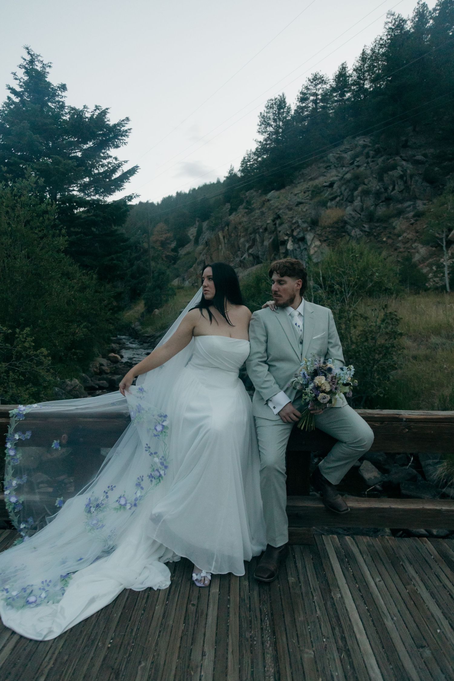 Bride and groom pose on rustic wooden bridge with flowing embroidered veil and mountain backdrop.