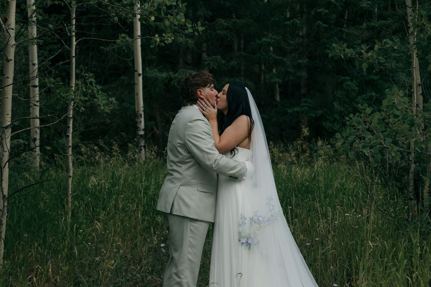 Bride and groom share a romantic first kiss surrounded by tall trees in a forest wedding.