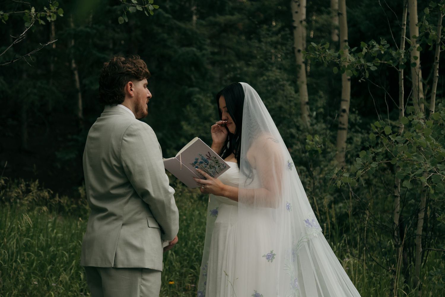 Bride reading wedding vows from a book during an outdoor forest ceremony, wearing a white gown and veil with groom in gray suit.