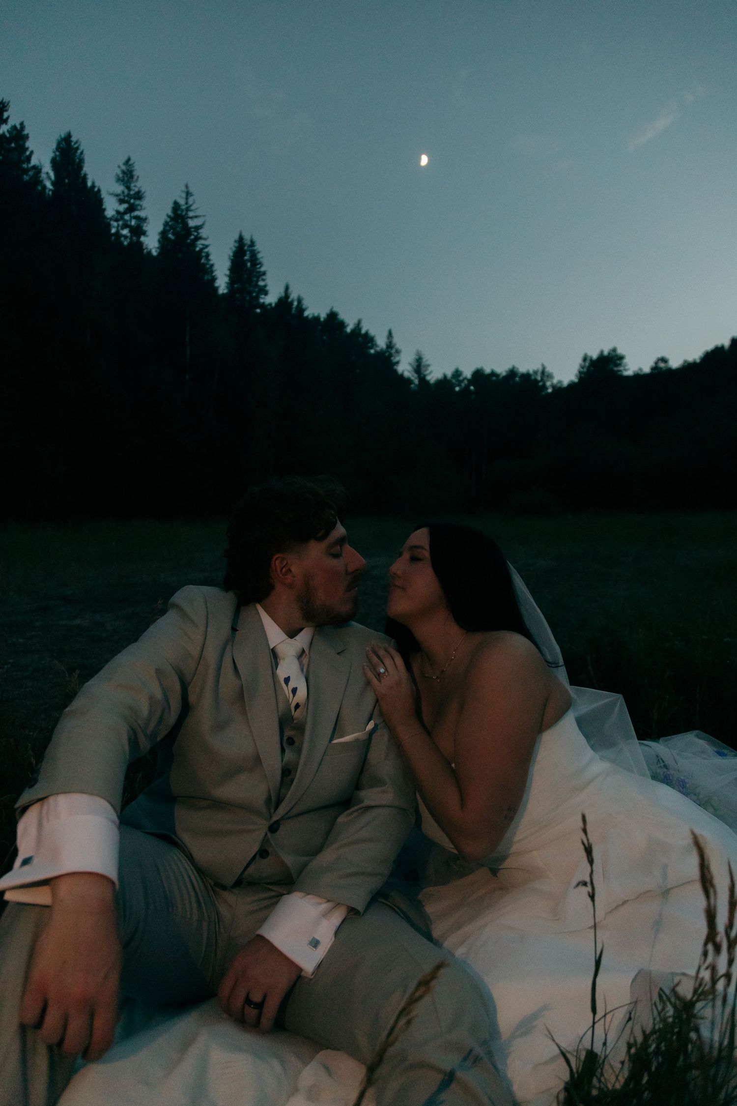 A bride and groom share a romantic moment sitting in a field under a crescent moon at dusk.