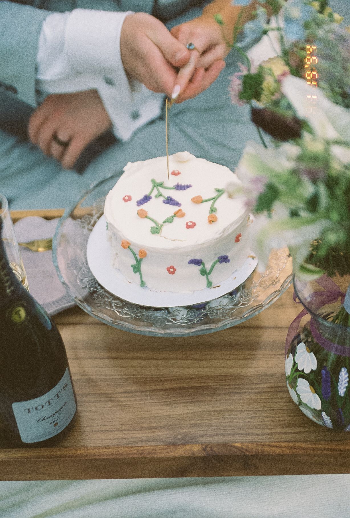 A small white wedding cake decorated with colorful embroidered flowers is lit with a sparkler beside champagne and florals.