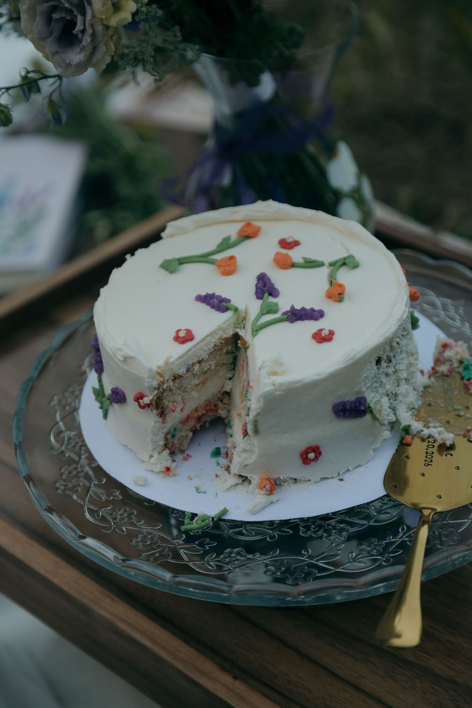 White wedding cake decorated with colorful pressed flowers, partially sliced on glass cake stand.
