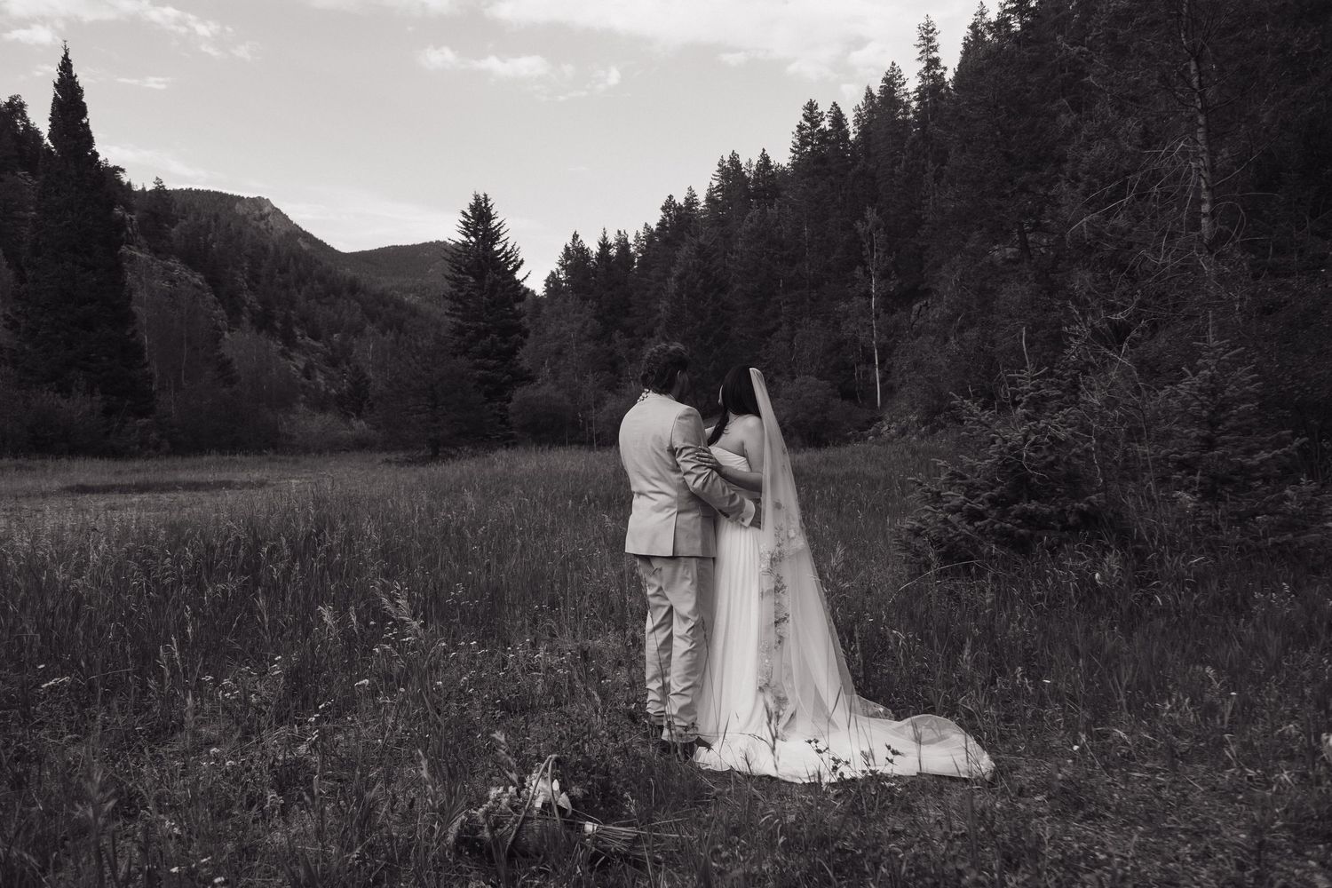 Black and white photo of bride and groom embracing in a mountain meadow surrounded by pine trees.