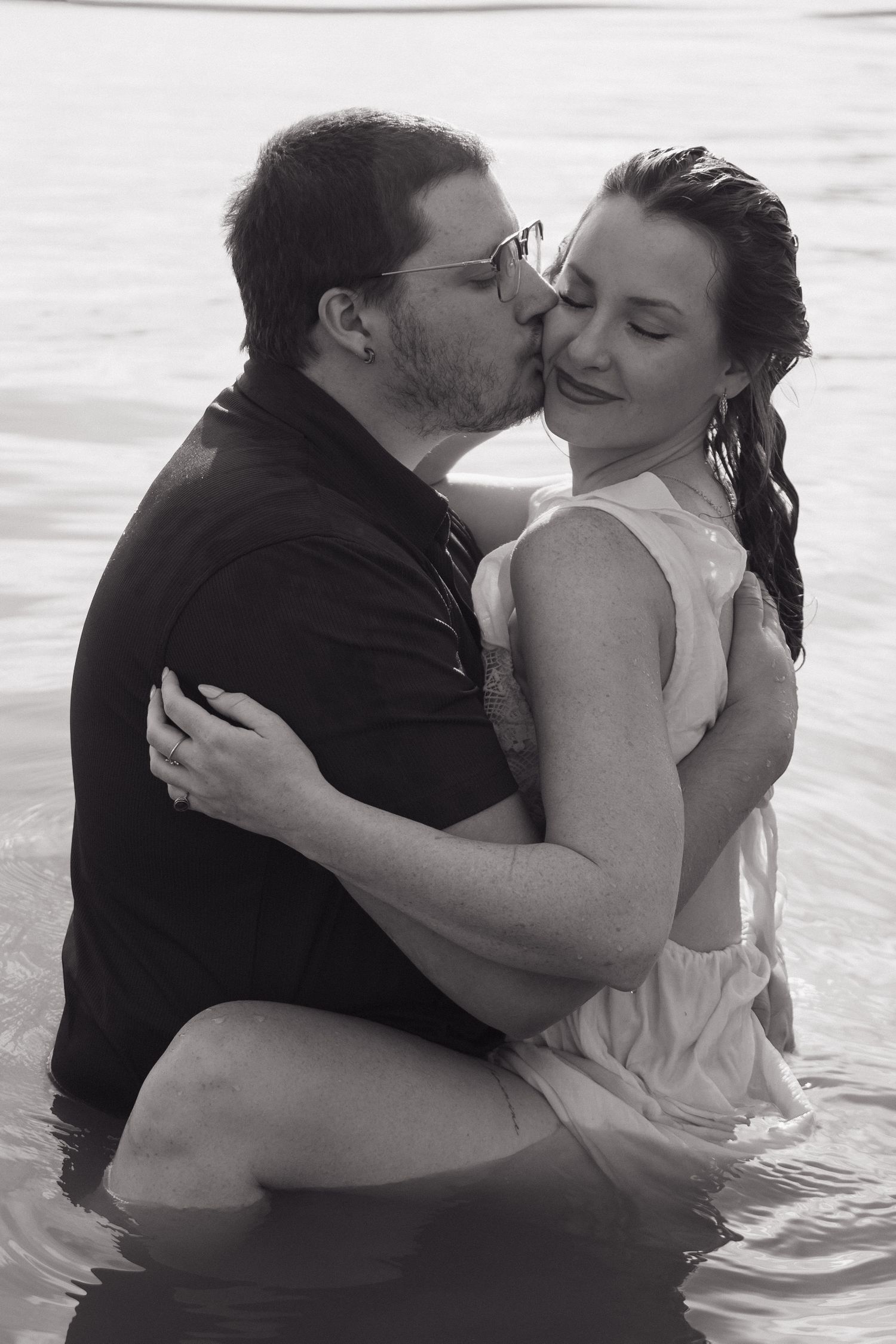 Black and white photo of man kissing woman's cheek while holding her in a lake, both fully clothed.
