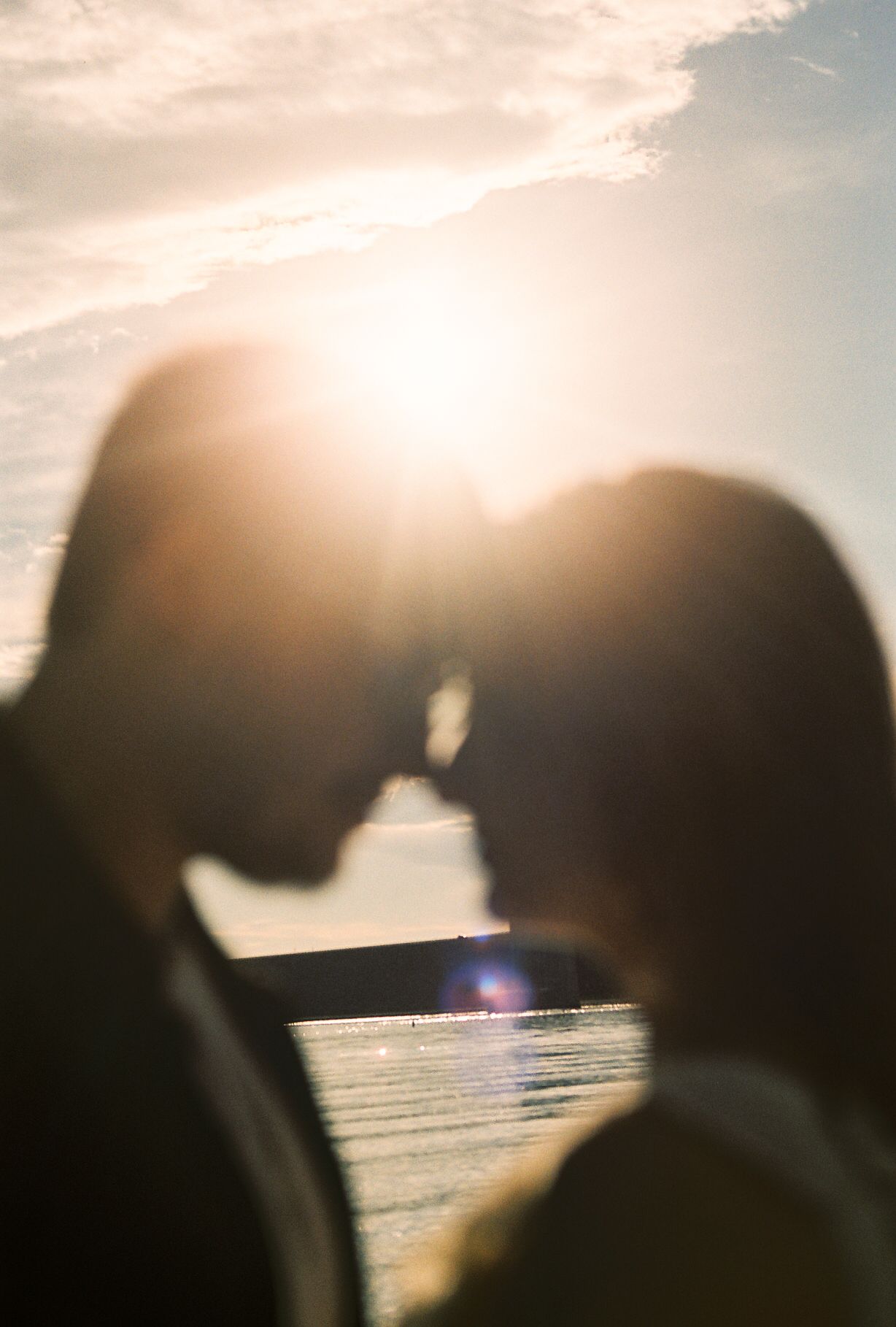 Silhouette of couple about to kiss backlit by brilliant golden sunset over calm water.