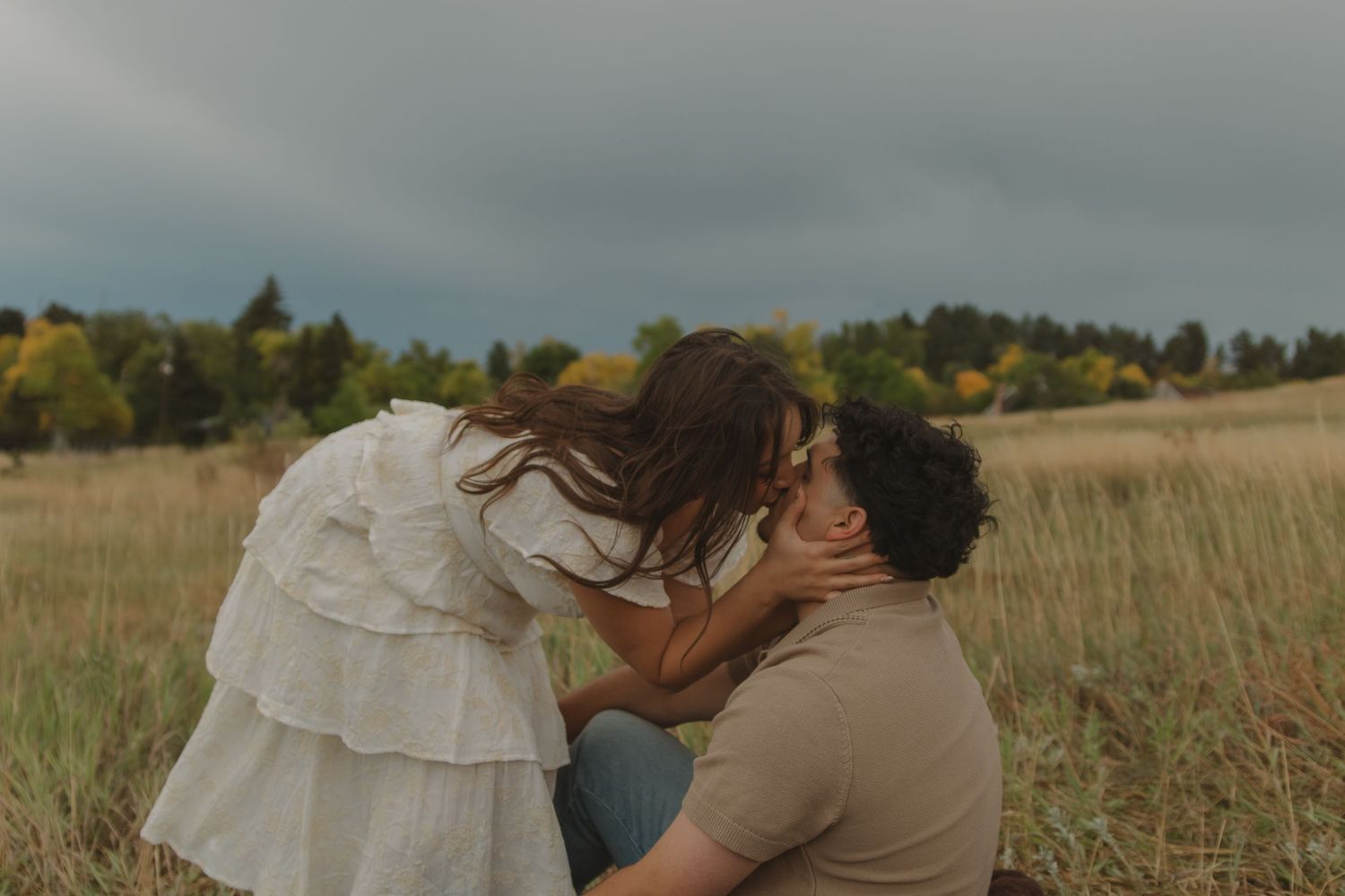 Woman leaning down to kiss man sitting in field under dramatic stormy sky.