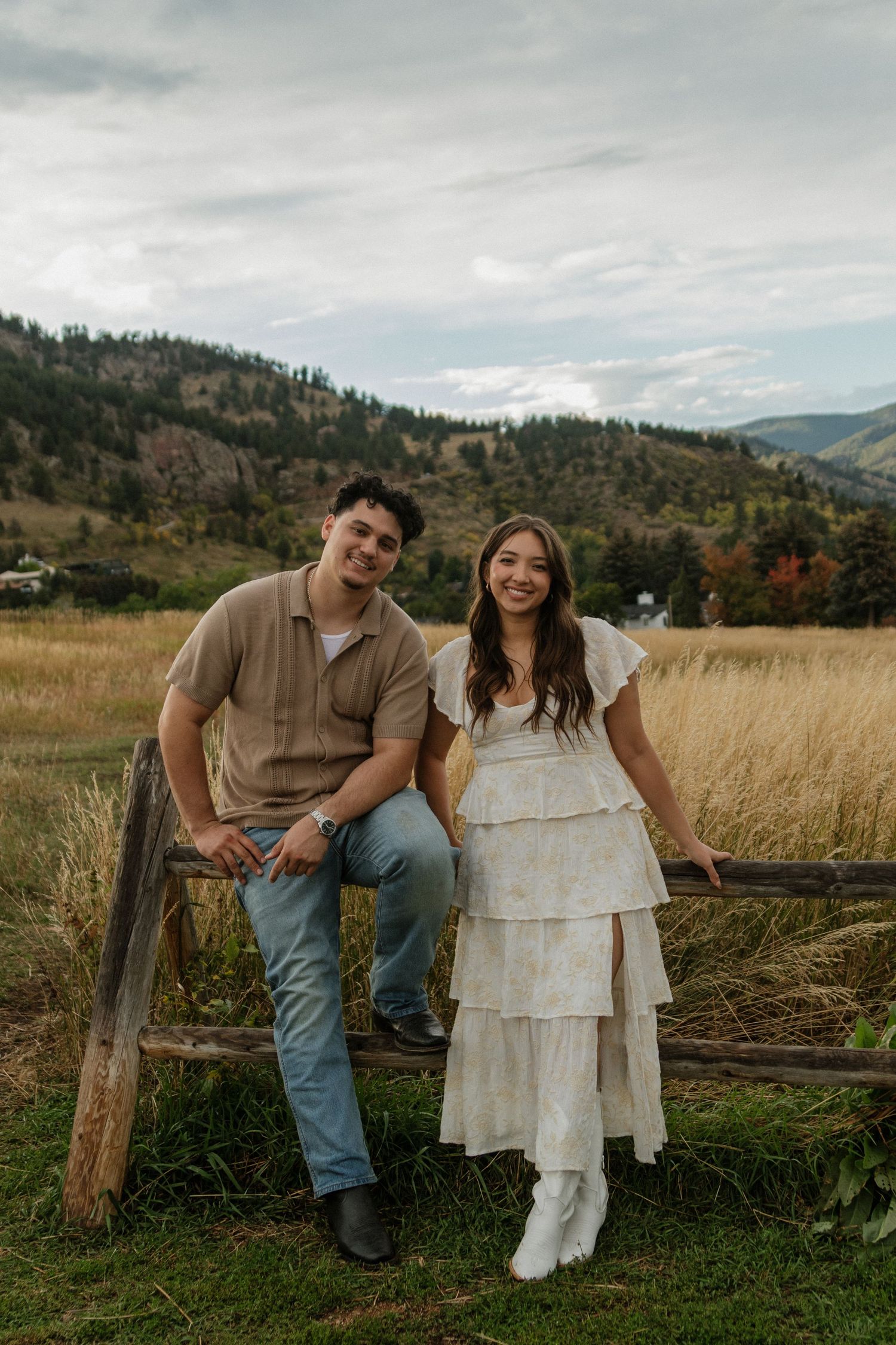 Smiling couple posing on a rustic wooden fence with golden fields and mountains behind.