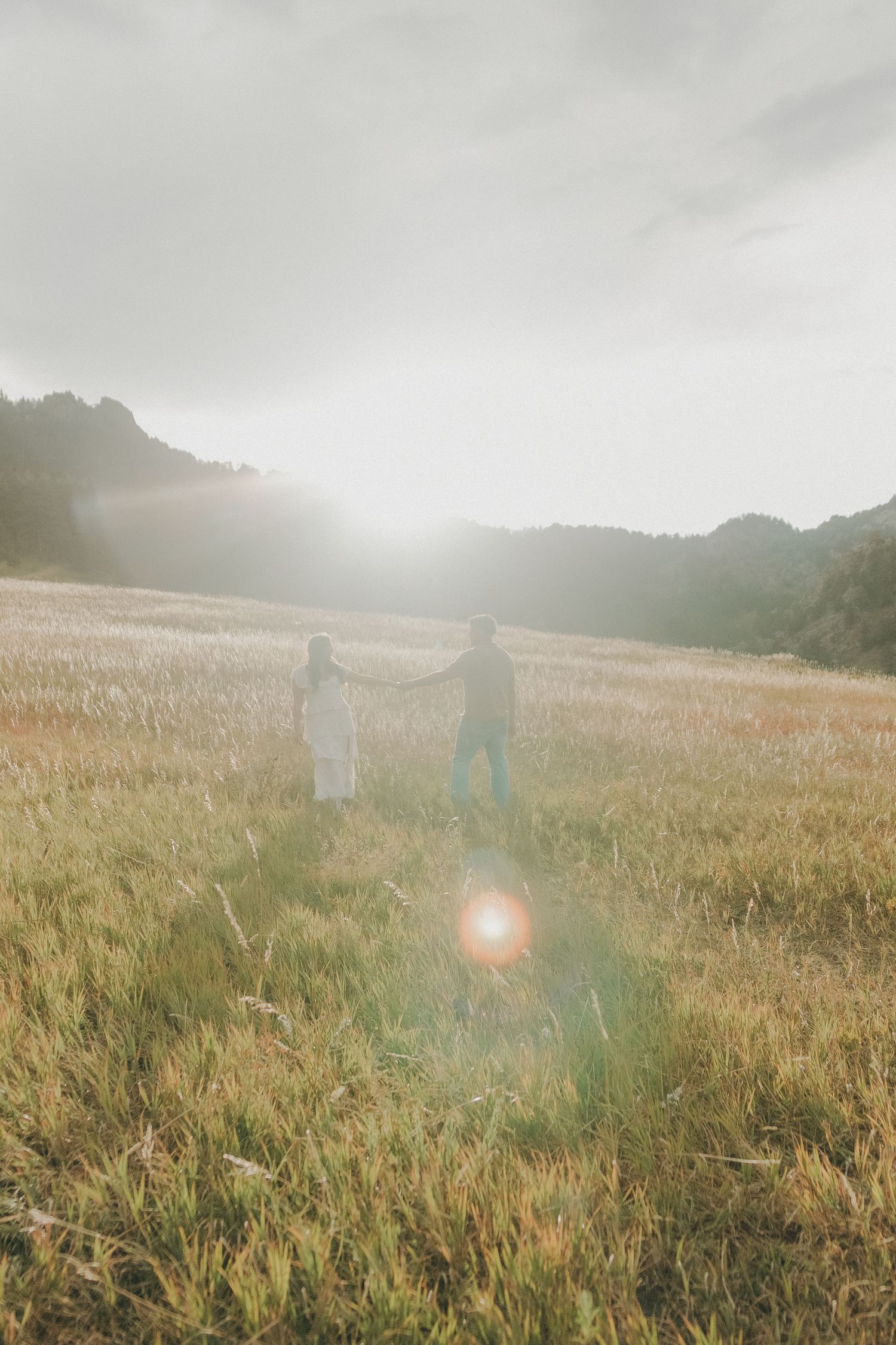 Couple holding hands walking through golden field with soft sunlight and misty mountains in background.