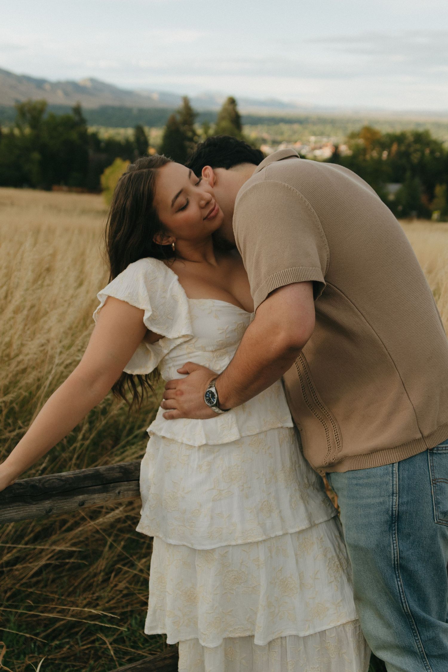 Romantic couple embracing in a golden field, man kissing woman in a white tiered dress at sunset.