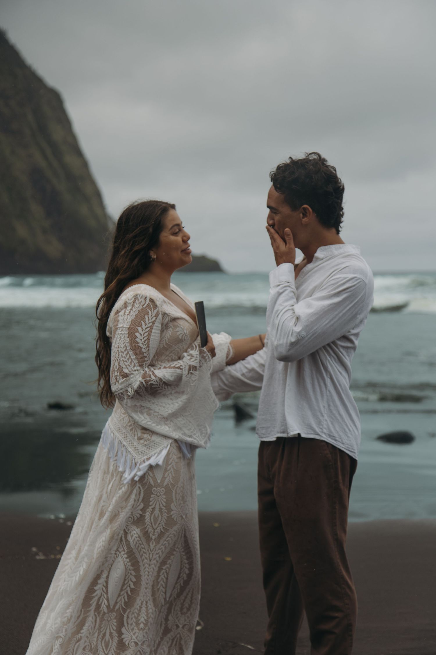 Bride in lace gown reads vows to groom on black sand beach with dramatic cliffs and ocean backdrop.