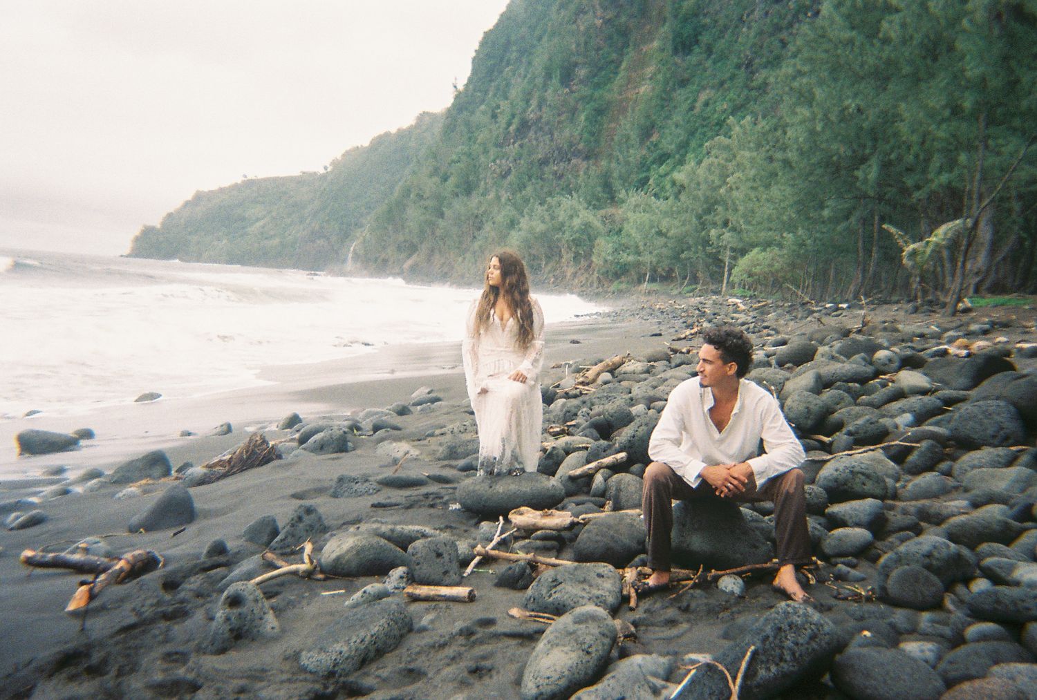 Couple in white clothing on a rocky black sand beach with lush green cliffs and ocean waves in background.