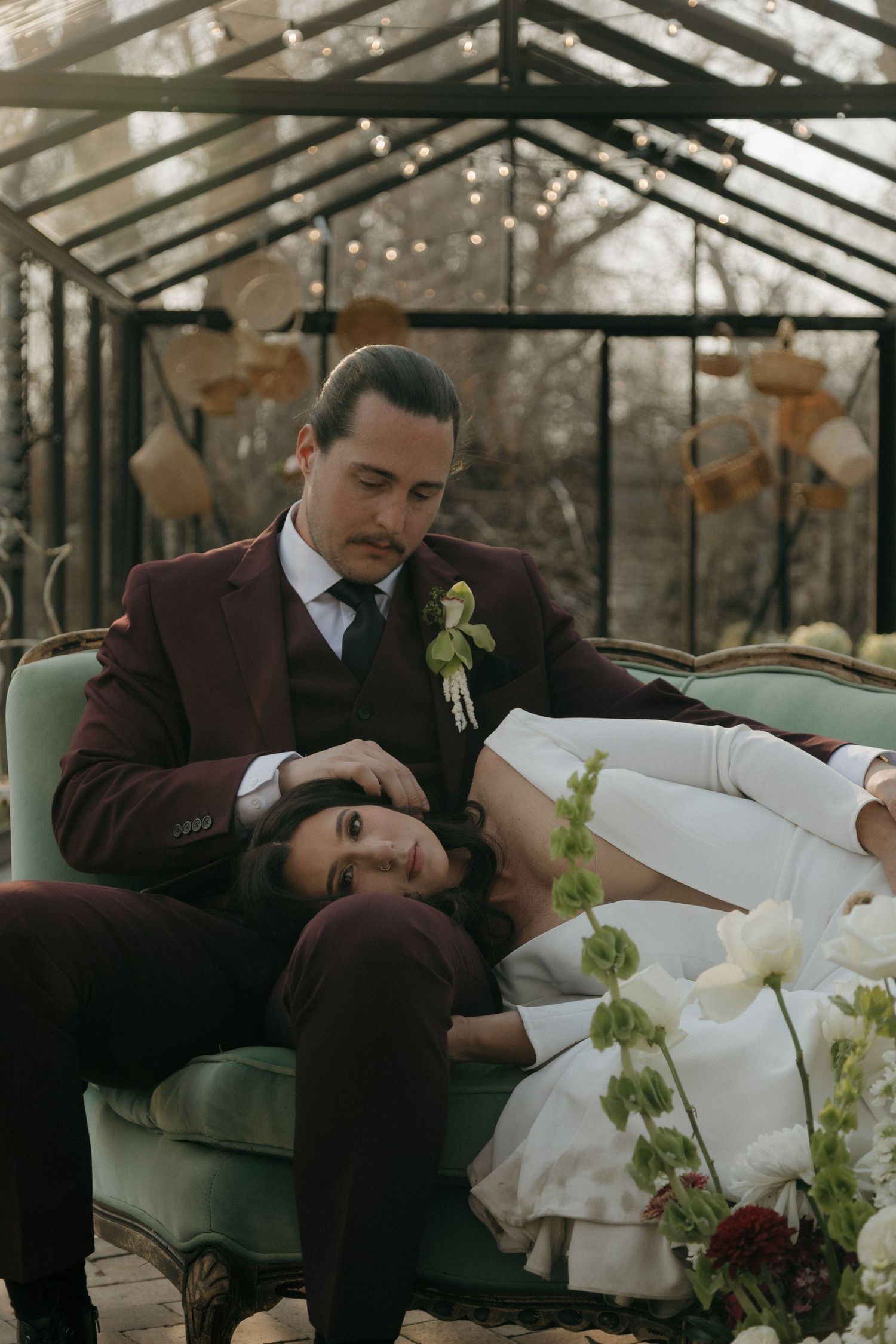 Groom in burgundy suit holds bride in white gown on vintage green sofa inside glass greenhouse.