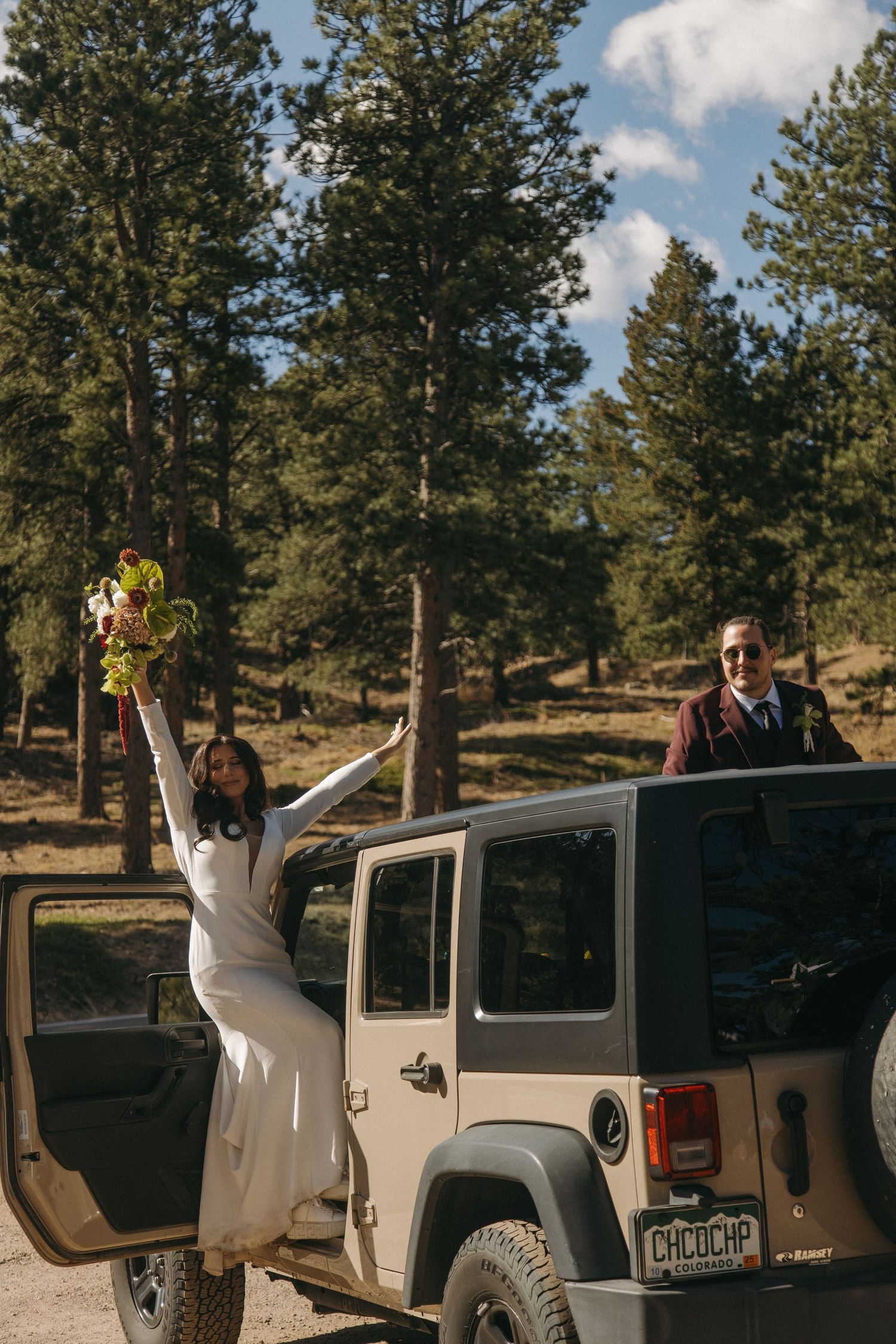 Joyful bride raises bouquet from Jeep door as groom stands through sunroof among pine trees.