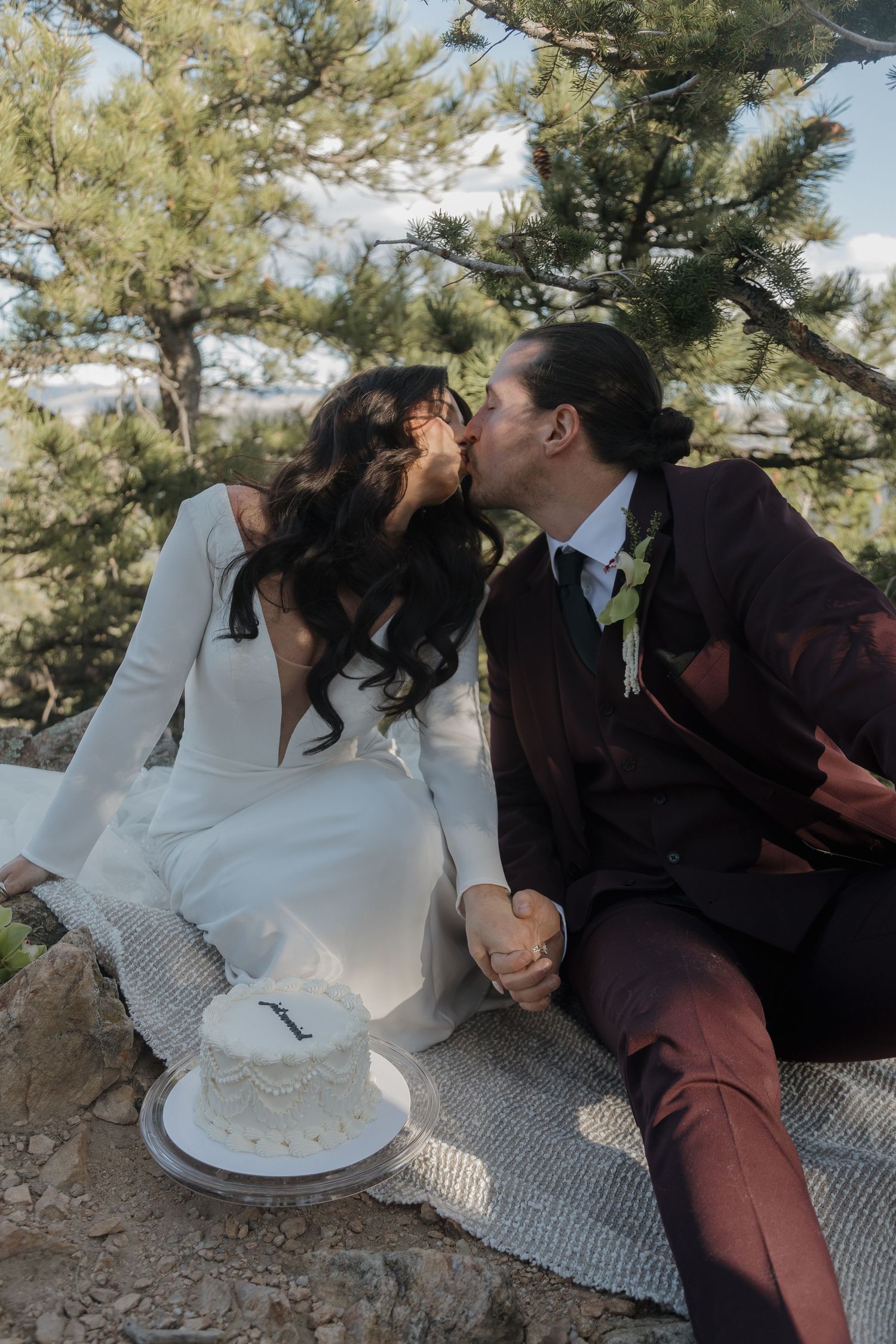 Bride and groom sharing a kiss while seated outdoors with a small white wedding cake, surrounded by pine trees.
