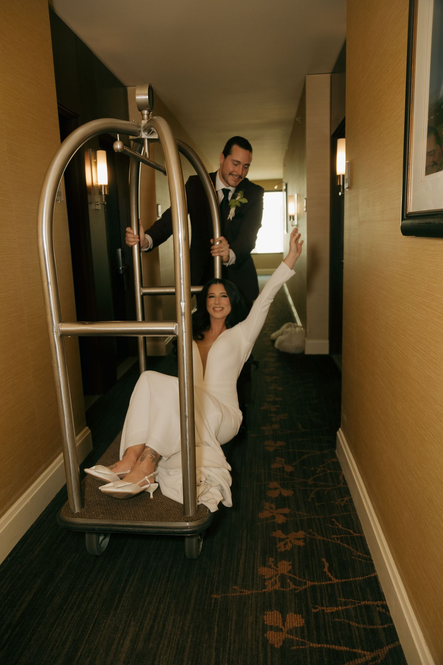 Bride sitting on hotel luggage cart while groom pushes her down hallway in playful wedding photo.