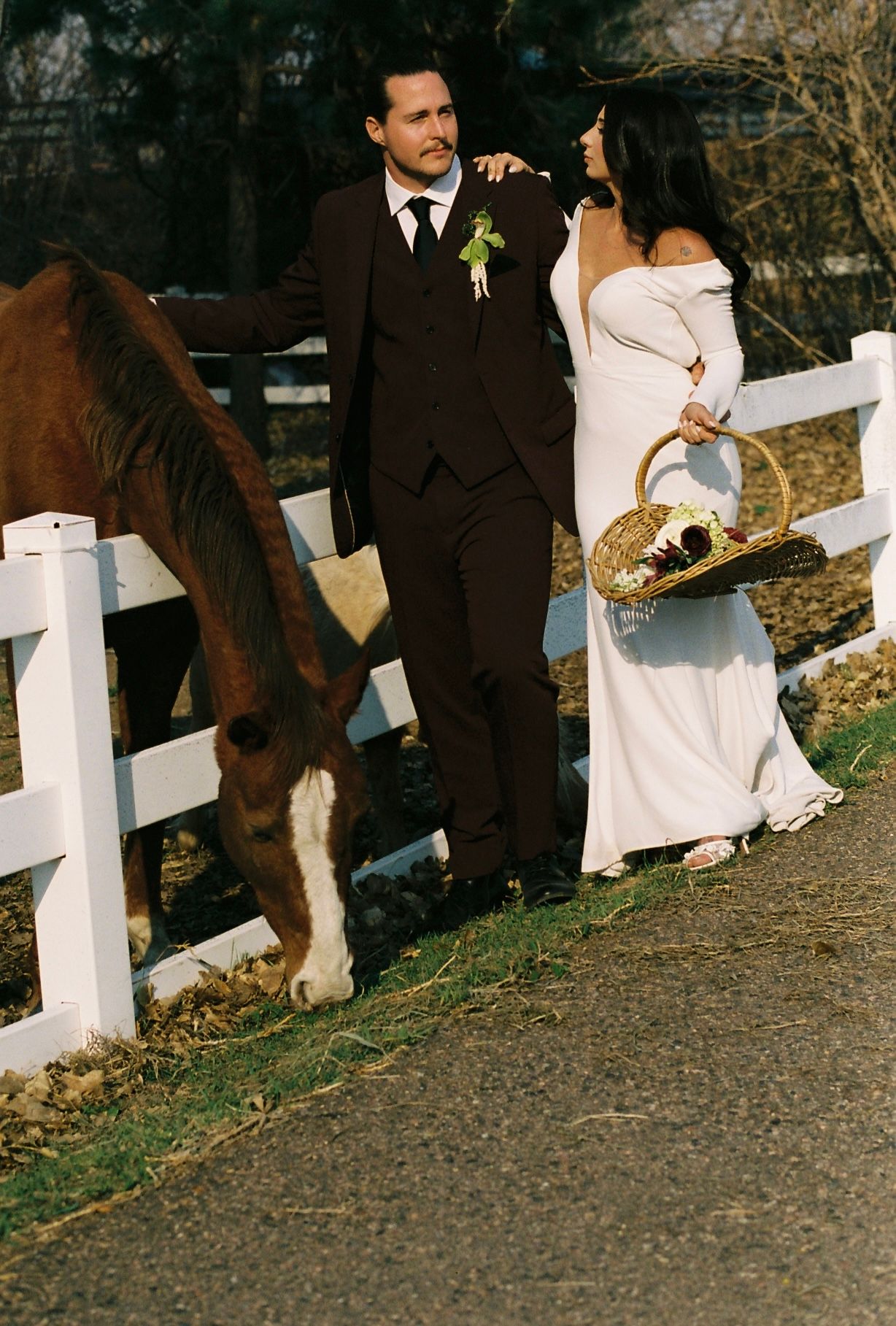 Bride and groom pose beside grazing chestnut horse at white fence farm in outdoor wedding portrait.