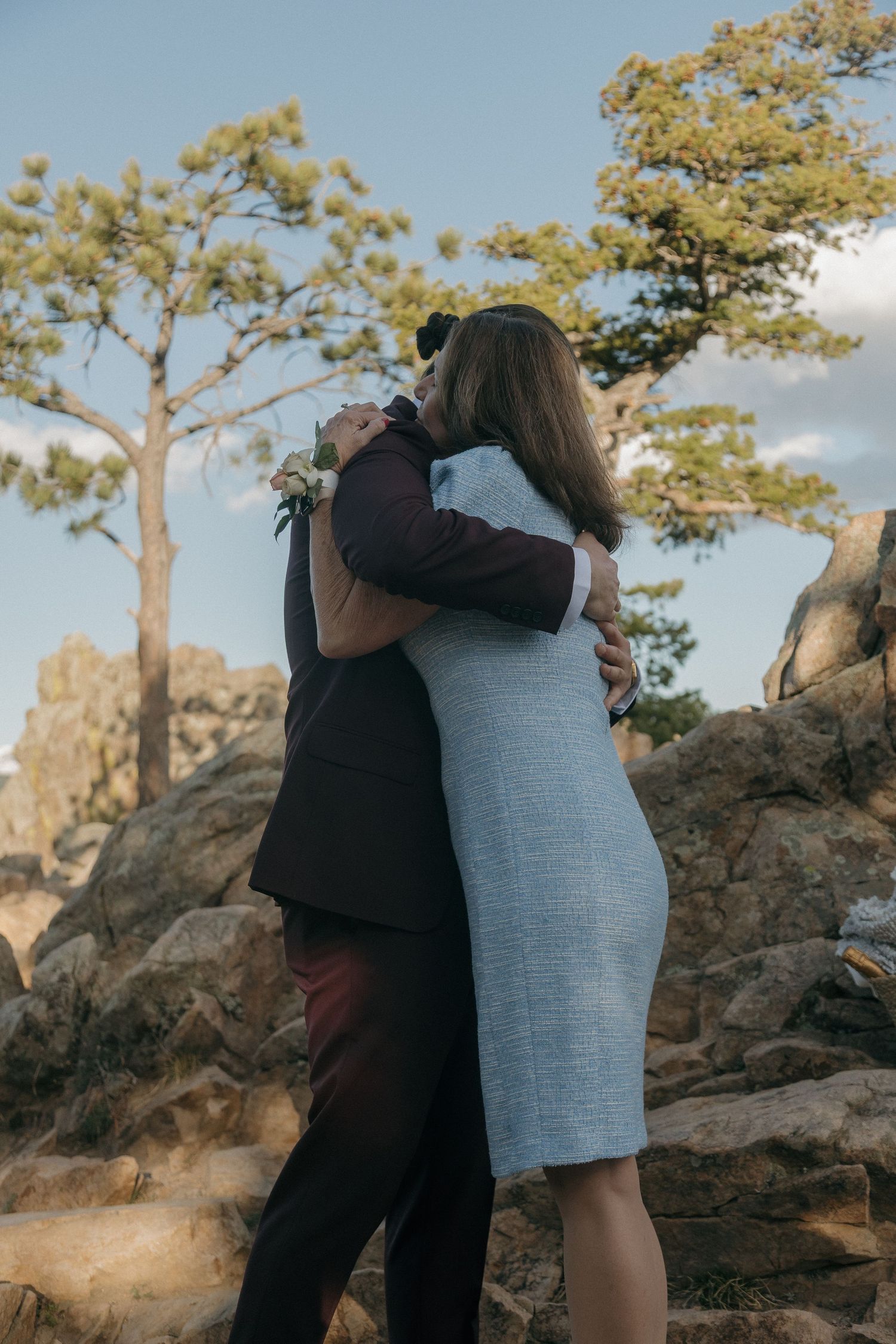 Groom in burgundy suit and guest in light blue dress share warm embrace at rocky outdoor wedding venue.