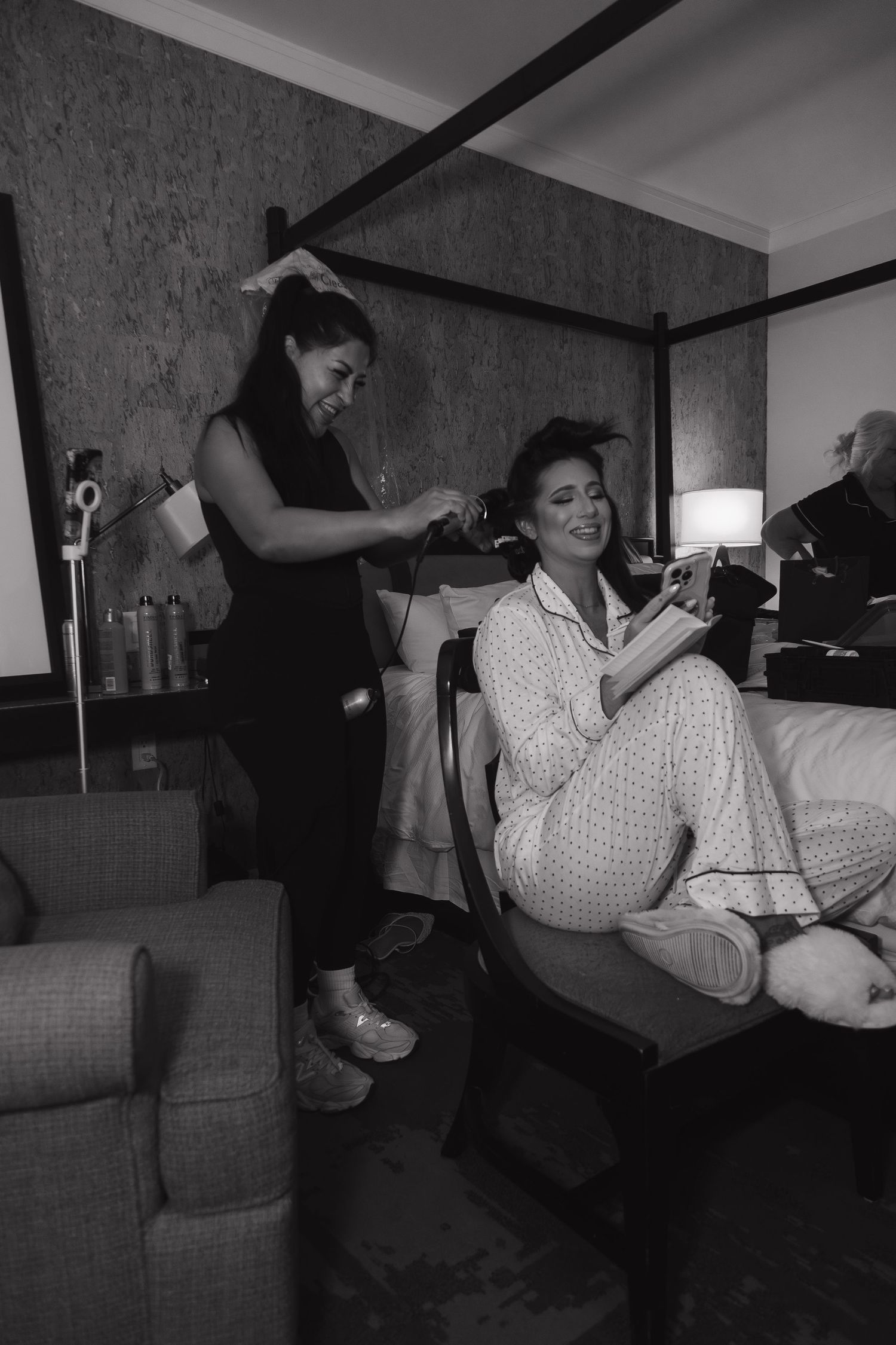 A hairstylist blow-dries a smiling woman's hair in a hotel room in black and white photo.