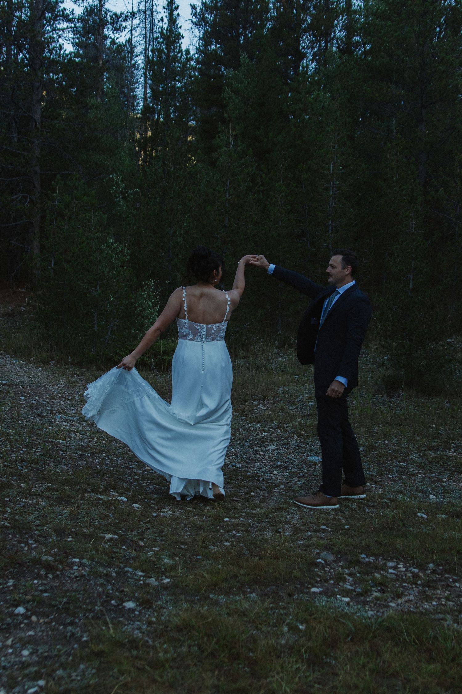 A bride in a white dress twirls on rocky terrain while her partner watches.