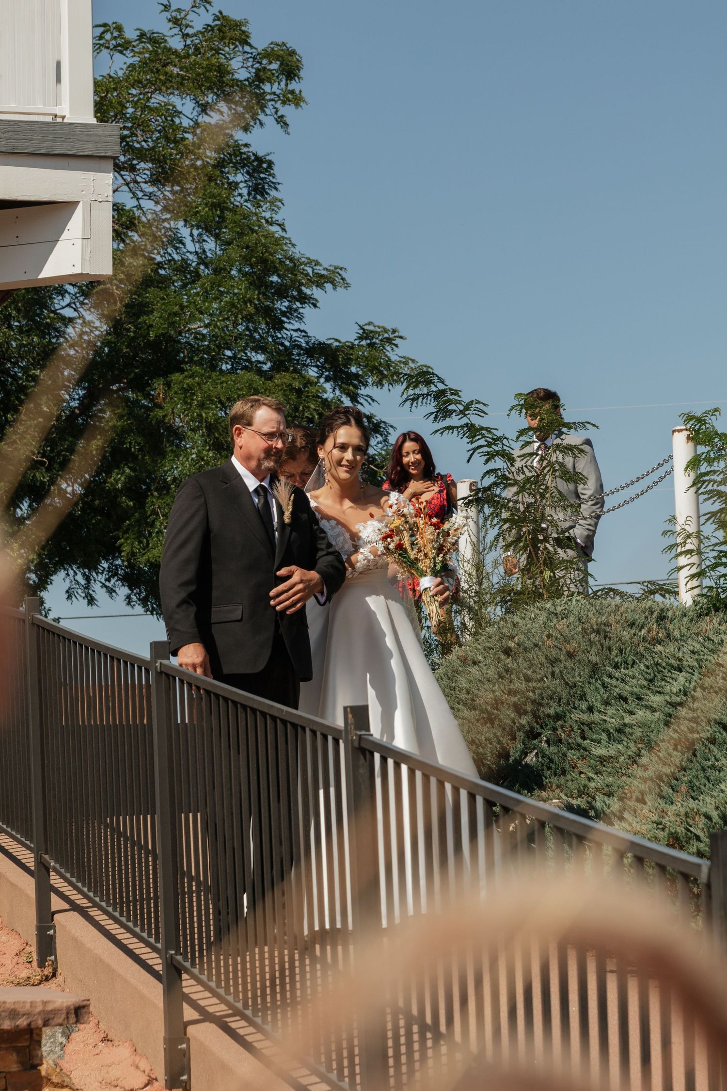 Wedding ceremony attendees line up along an outdoor railing with trees in the background.