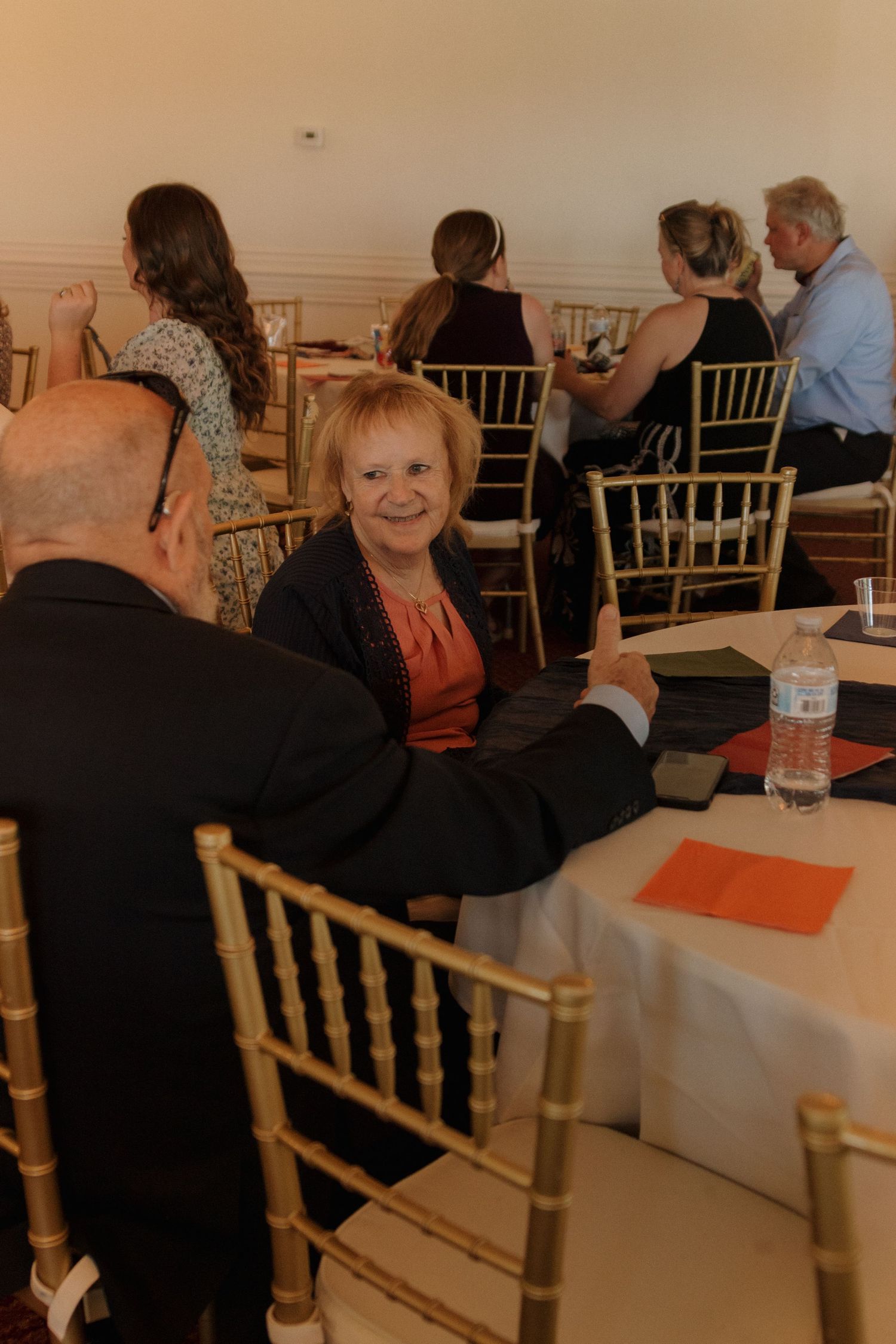 Wedding reception guests seated at tables with gold chiavari chairs and orange accents.
