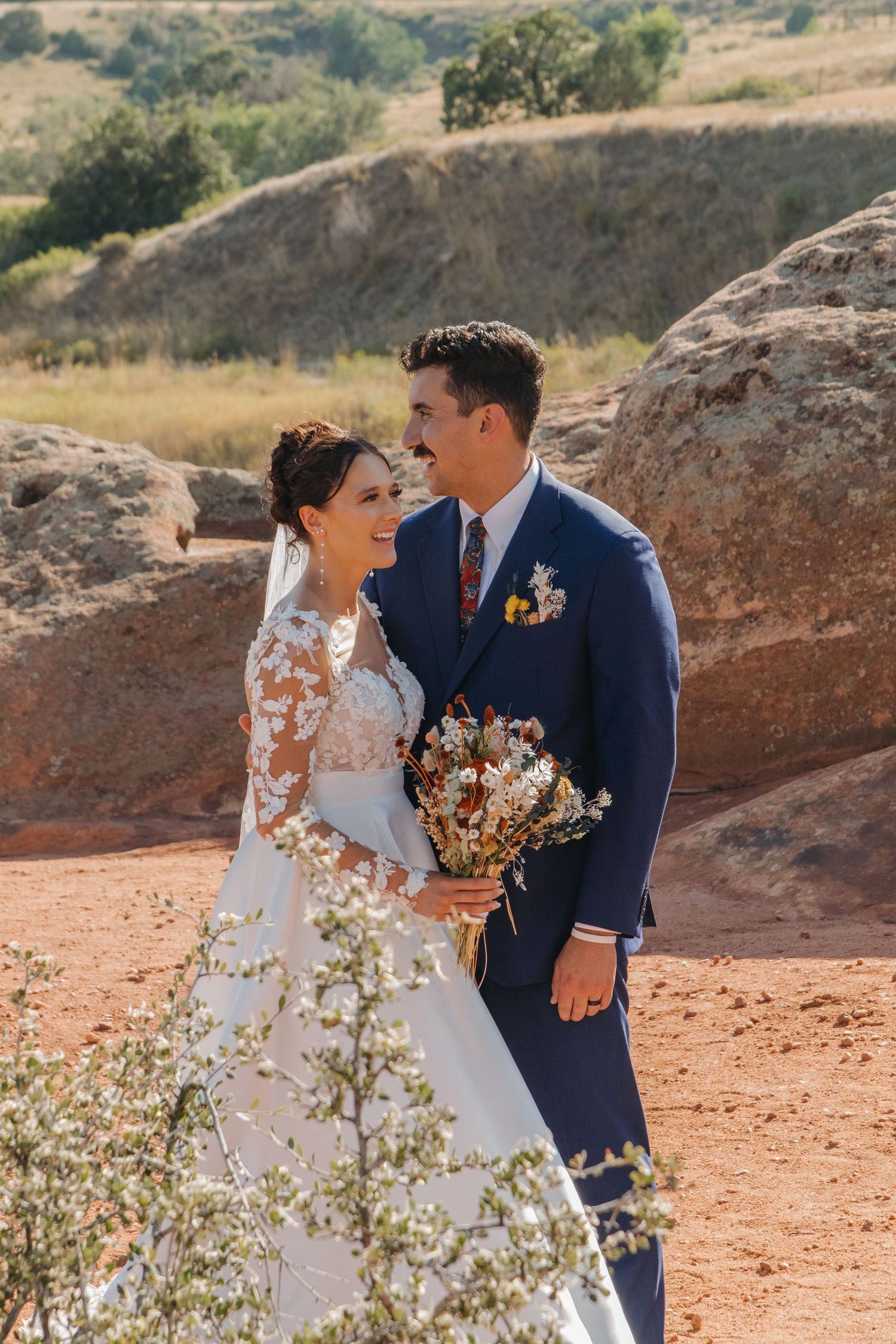 Newlyweds share an intimate moment on a red rock cliff with mountains in the background.