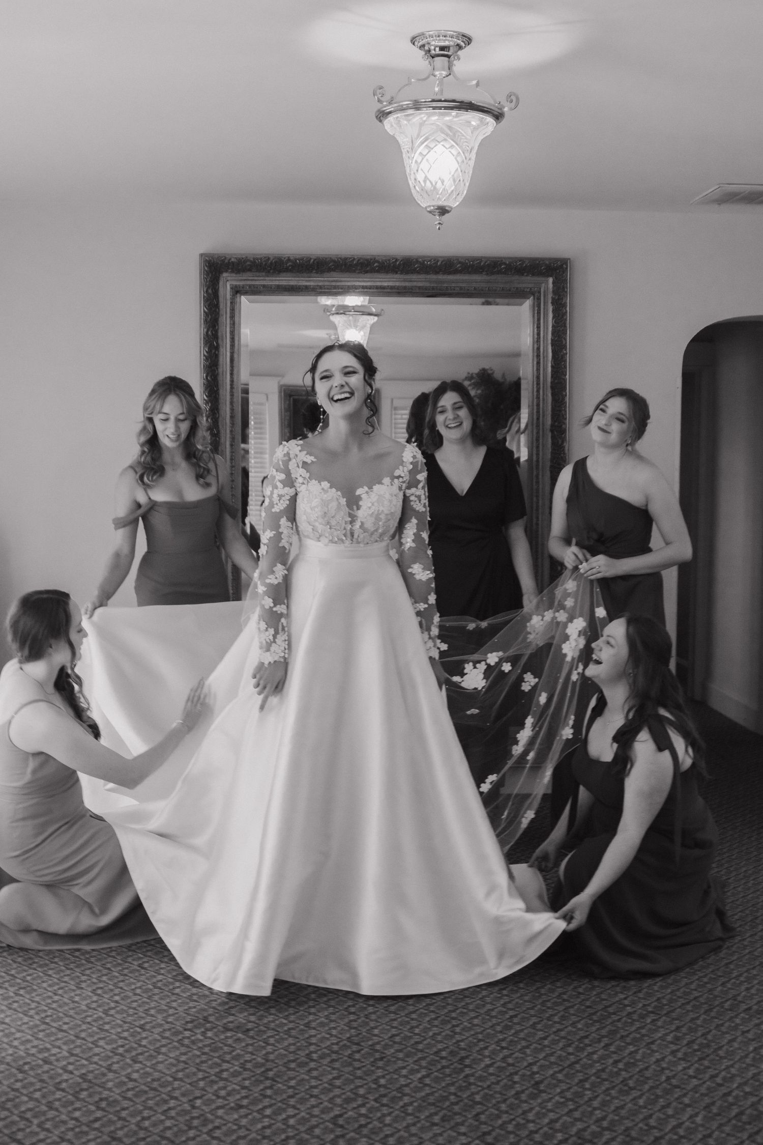 A wedding dress fitting moment captured in black and white with attendants helping arrange the gown.