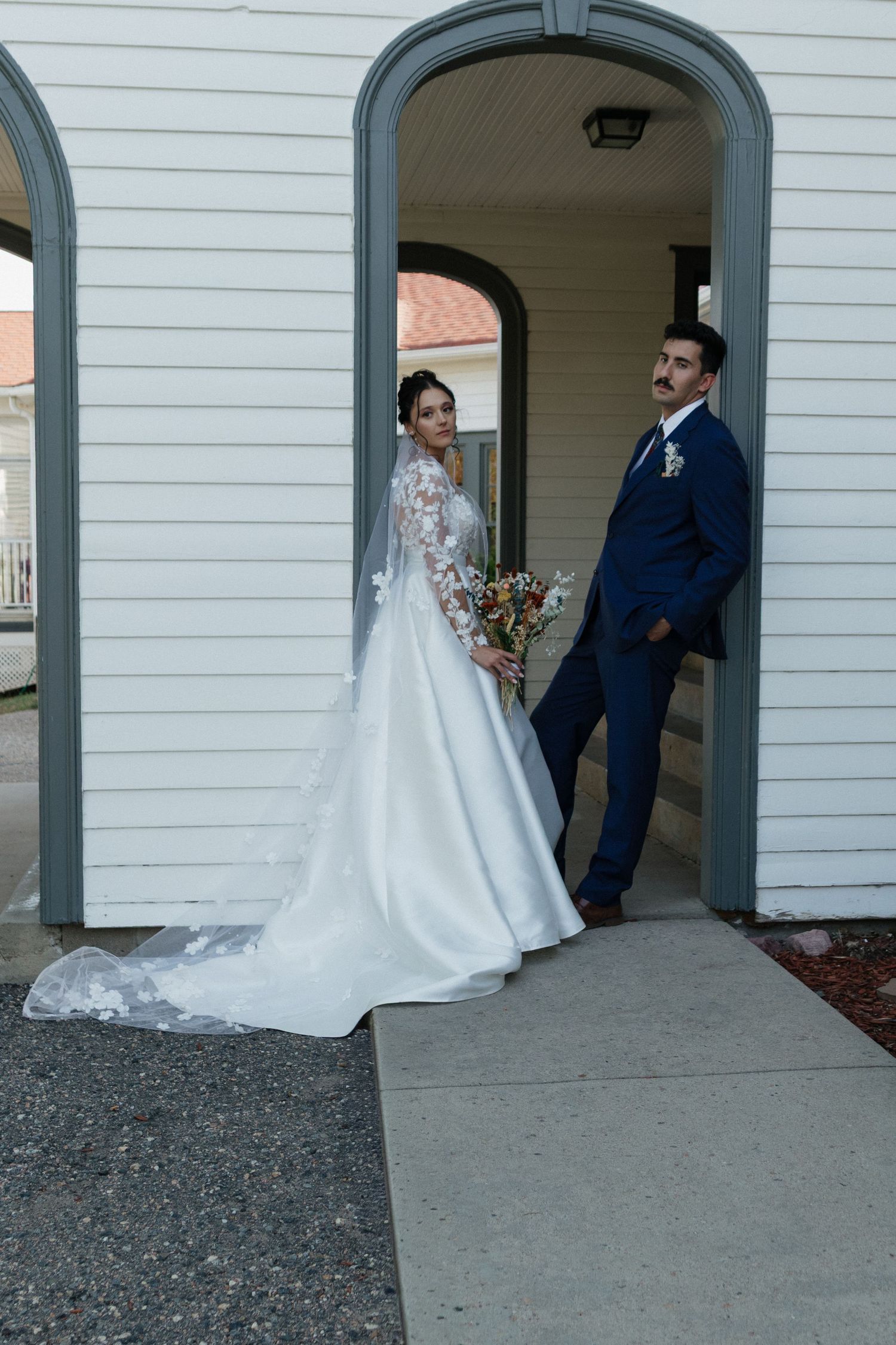 Newlyweds pose under white archways of a classic building in formal attire.