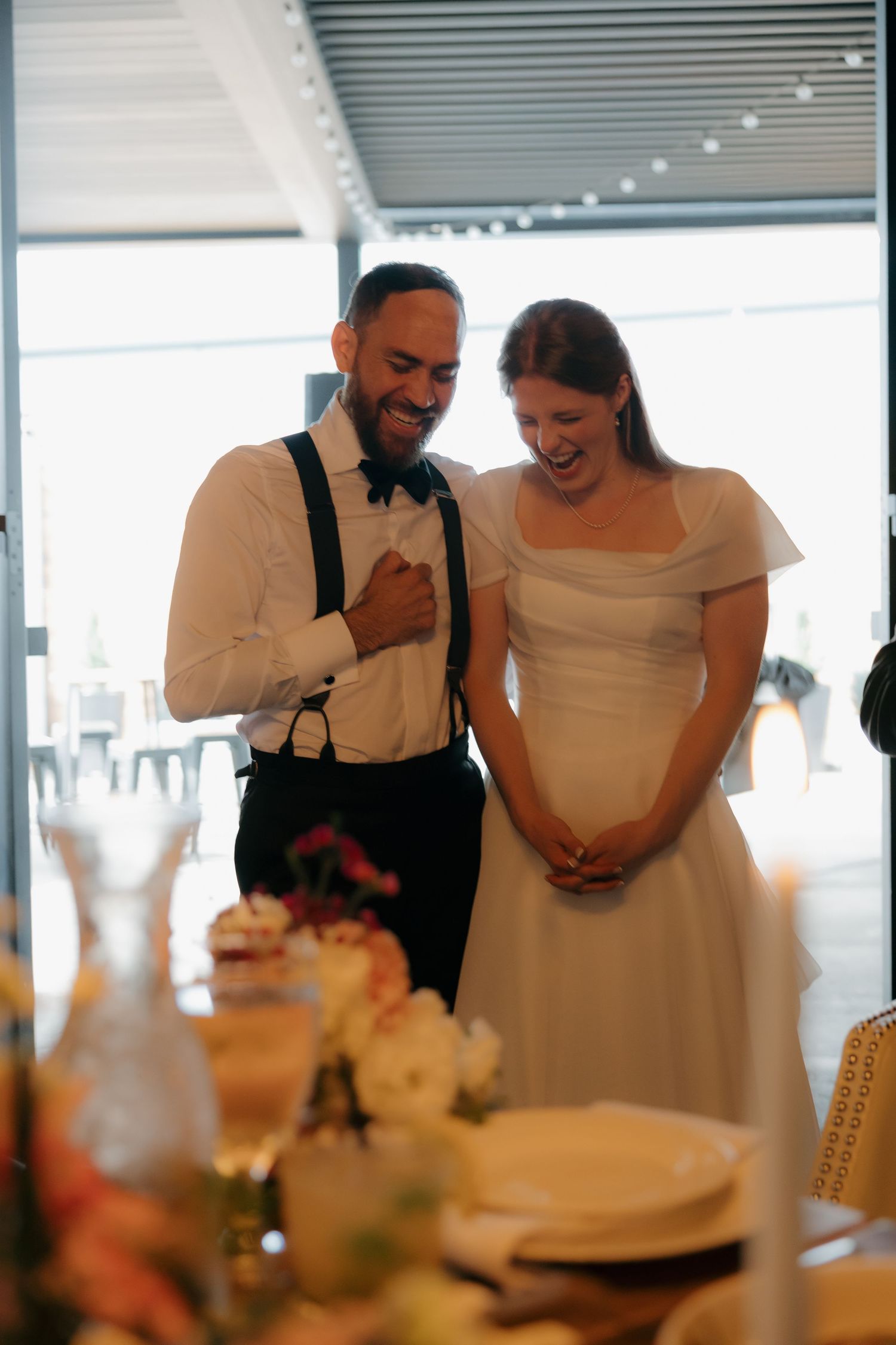 Laughing bride and groom in suspenders share joyful moment together beside floral-decorated wedding reception table.