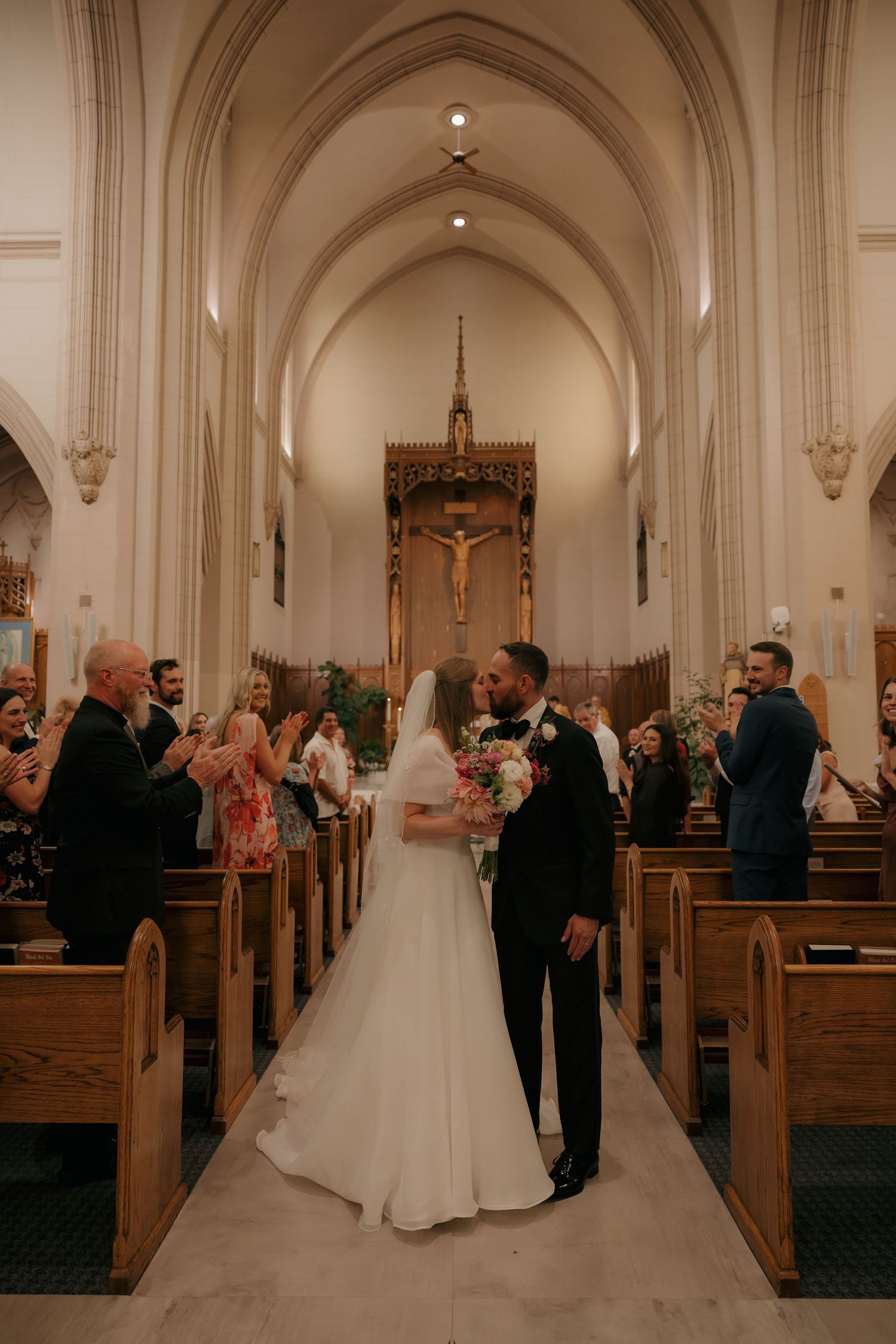 Newlyweds share first kiss as husband and wife in church aisle after wedding ceremony.