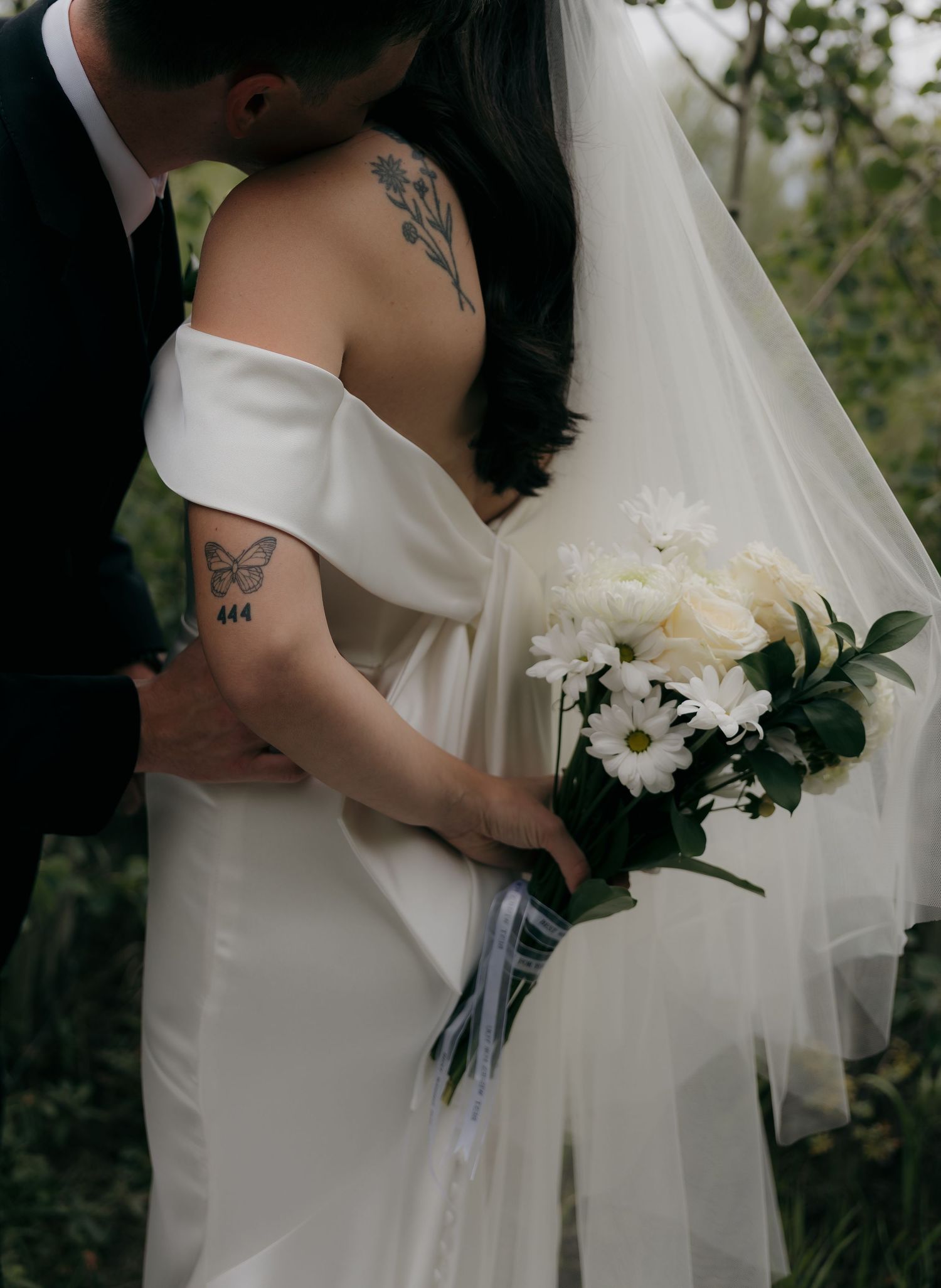 Groom kisses tattooed bride in off-shoulder wedding gown while holding white bouquet in nature.
