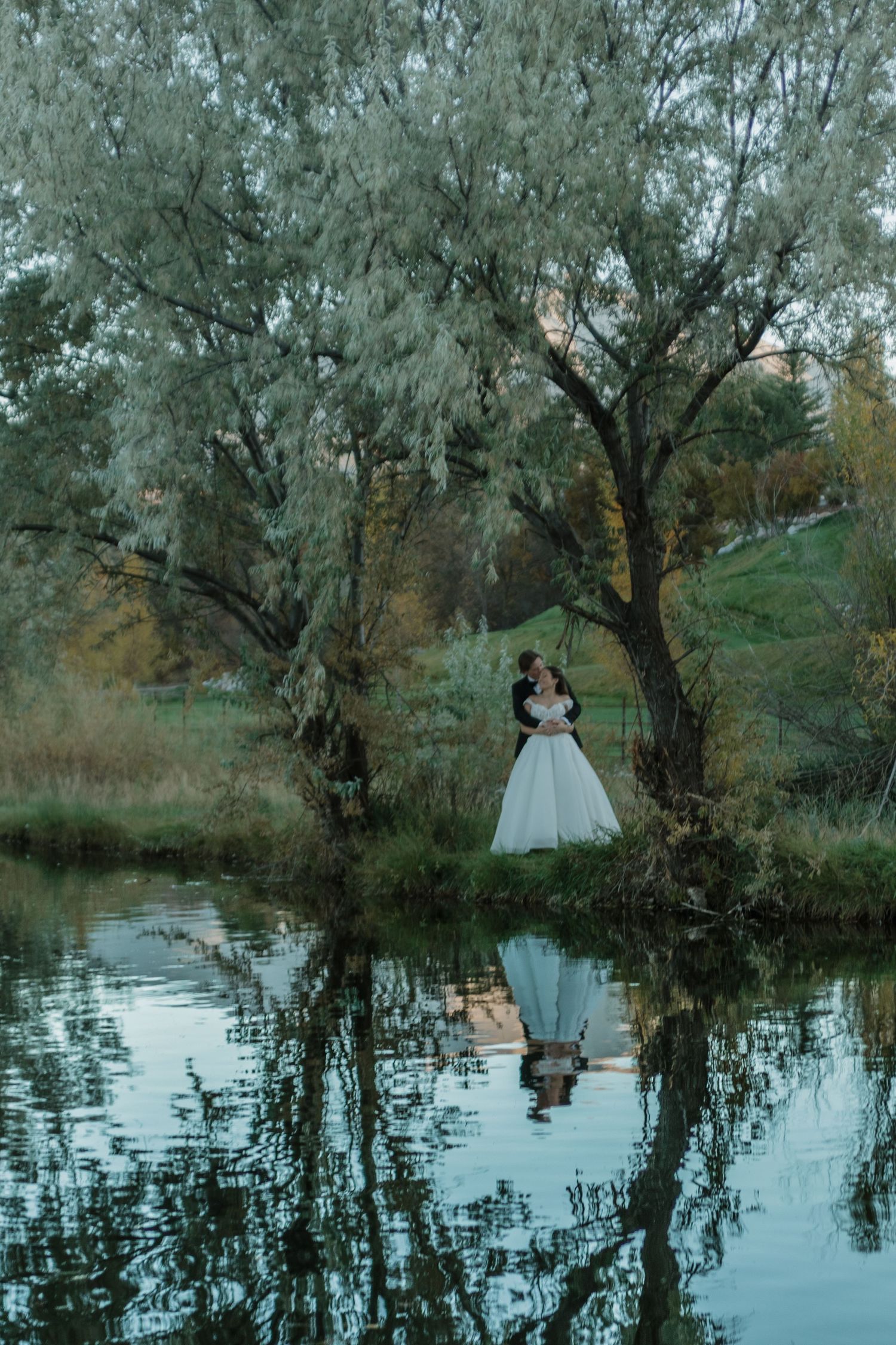 A romantic wedding photo with reflections in a pond surrounded by willow trees at dusk.