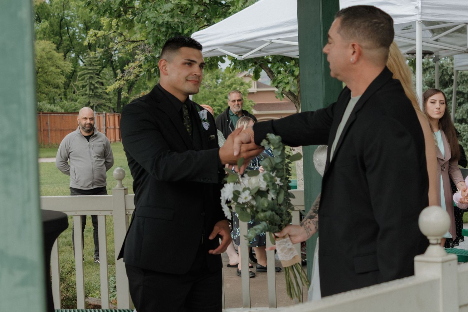 Wedding party members standing under green gazebo during outdoor ceremony.