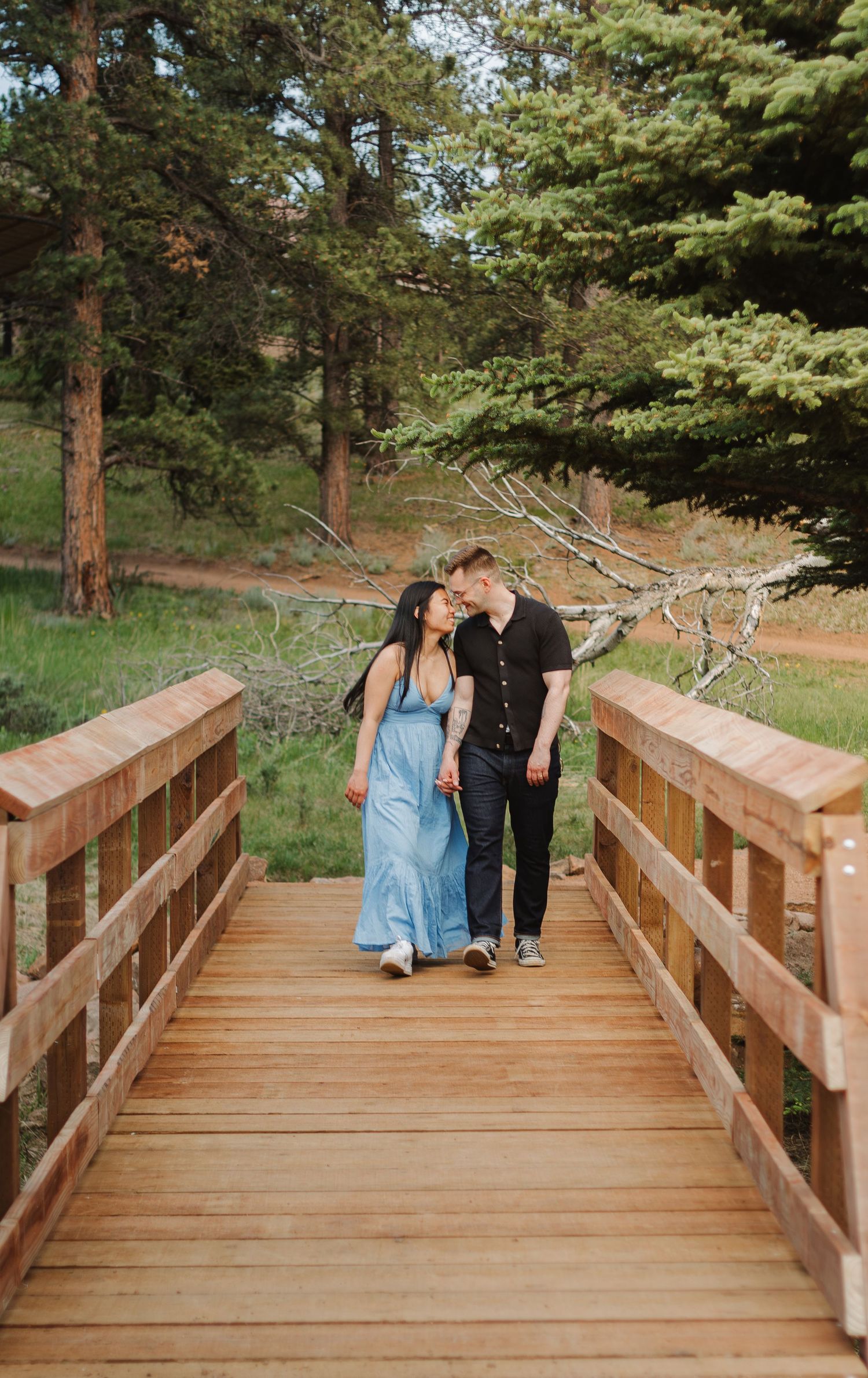 A couple walks together on a wooden bridge surrounded by evergreen trees.