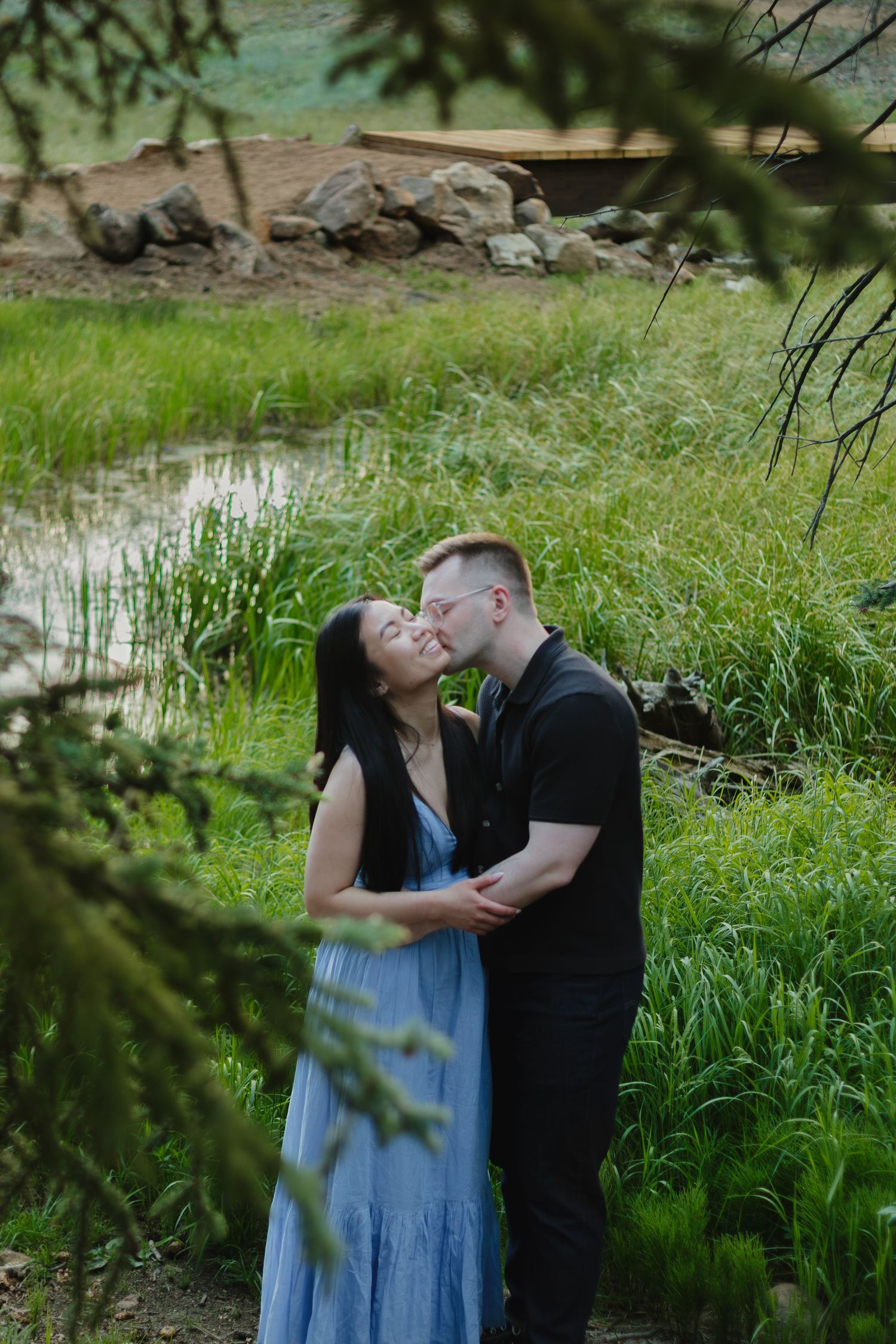 Couple sharing an intimate moment by a pond surrounded by green grass and pine trees.