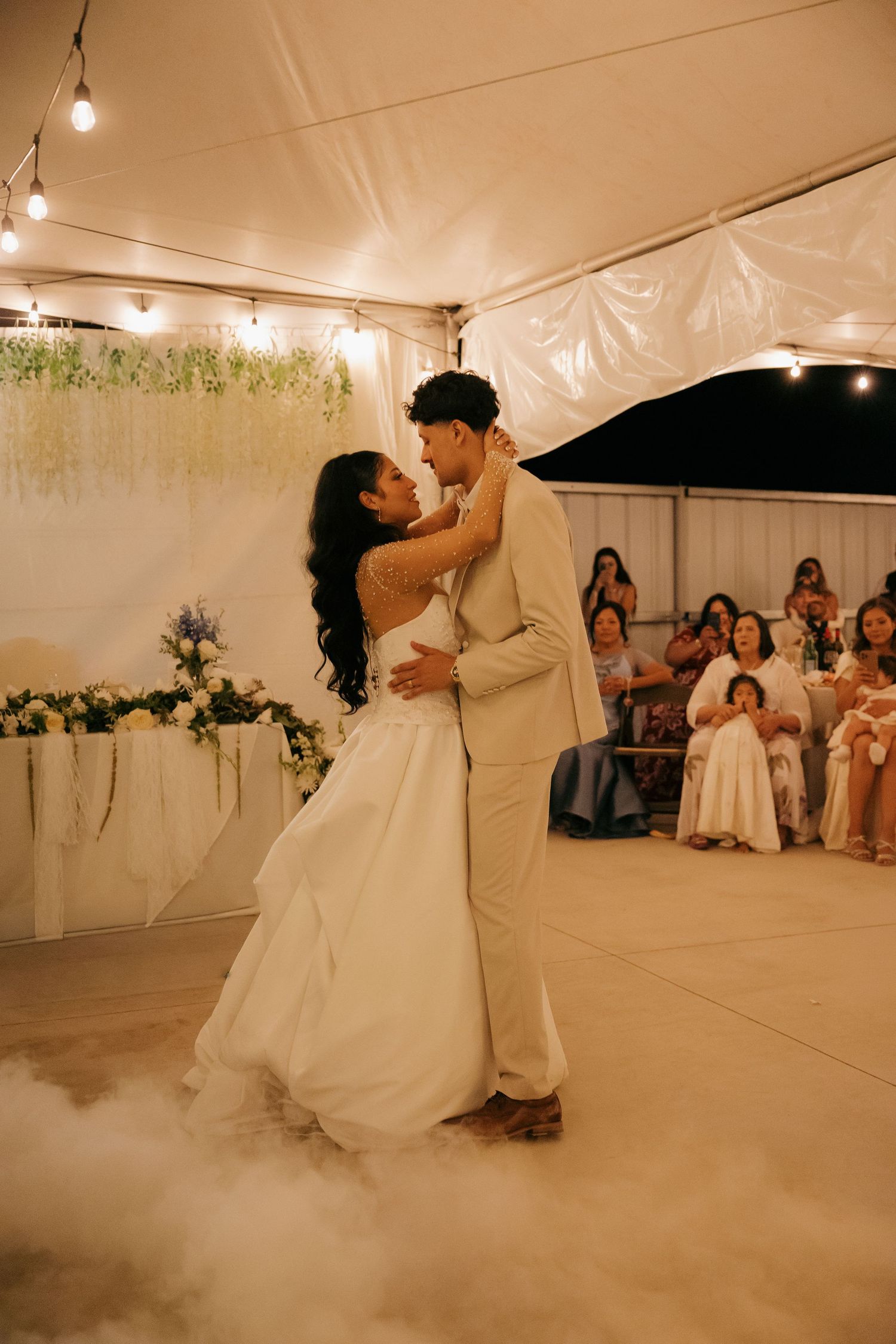 Bride and groom share romantic first dance surrounded by fog and floral decor at reception.
