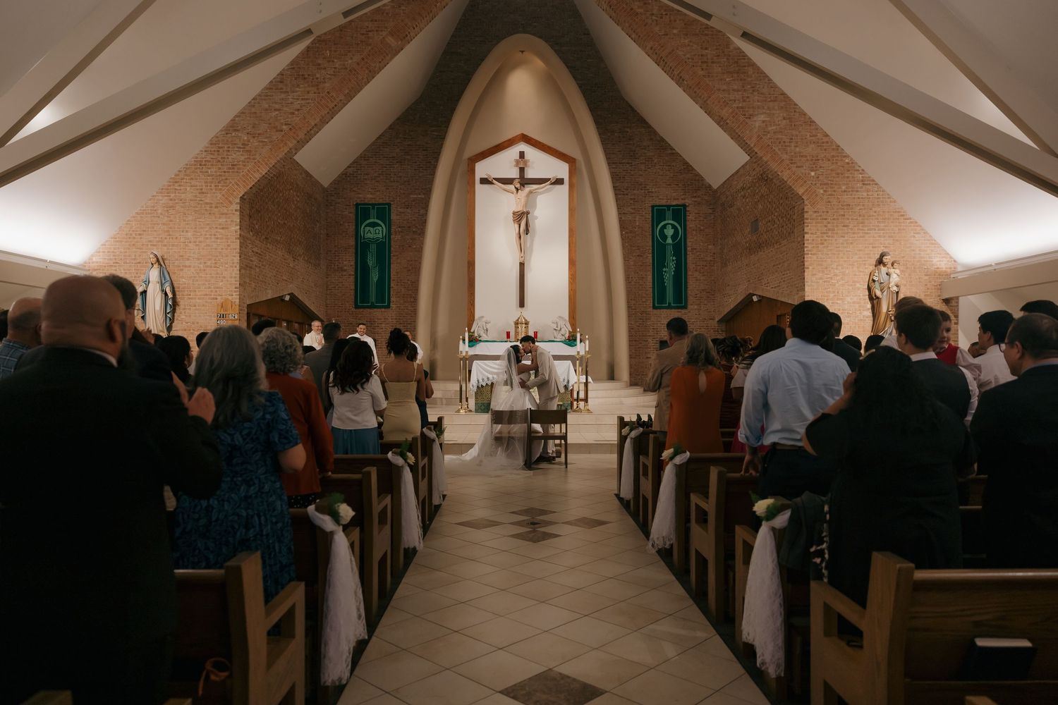Guests stand in wooden pews during a Catholic wedding ceremony in a chapel with a crucifix altar.