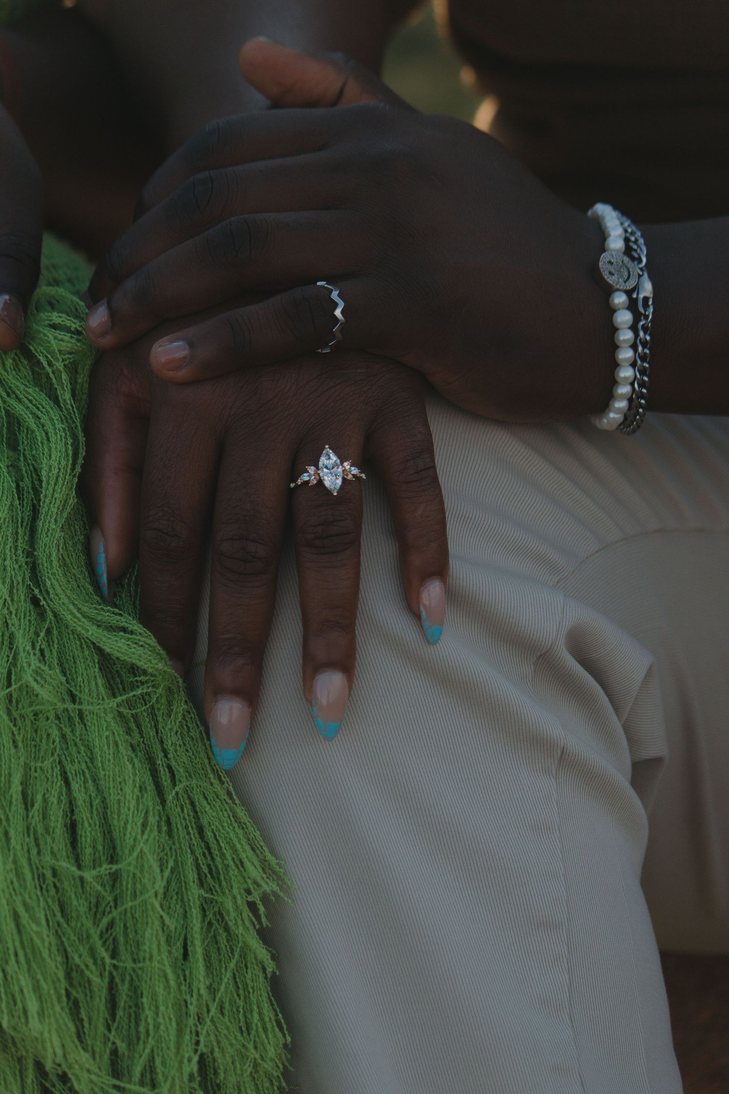 Close-up of hands showing off an engagement ring with green fabric and white clothing visible in the background.