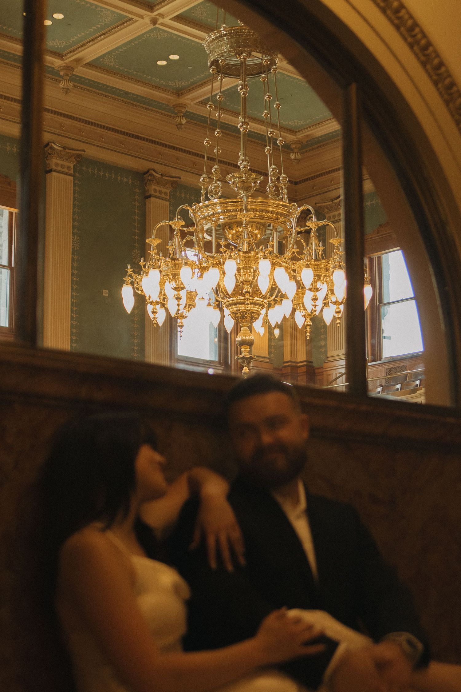 Couple sits beneath an elegant crystal chandelier in a grand historic building.